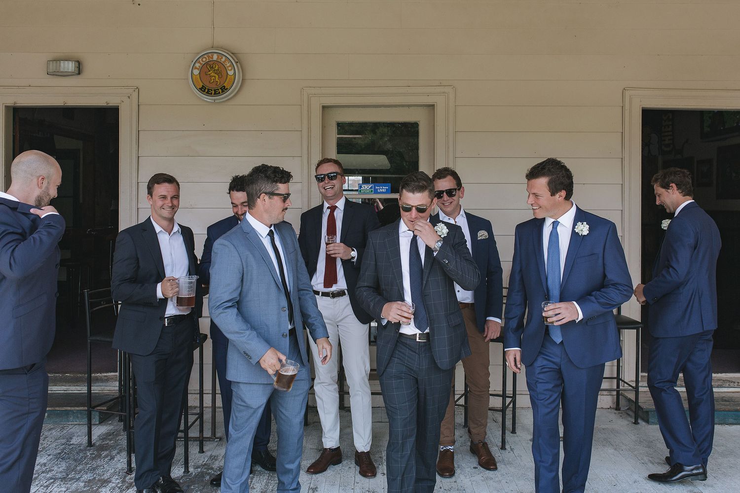 A group of groomsmen in suits and ties gather on a porch before a wedding ceremony.