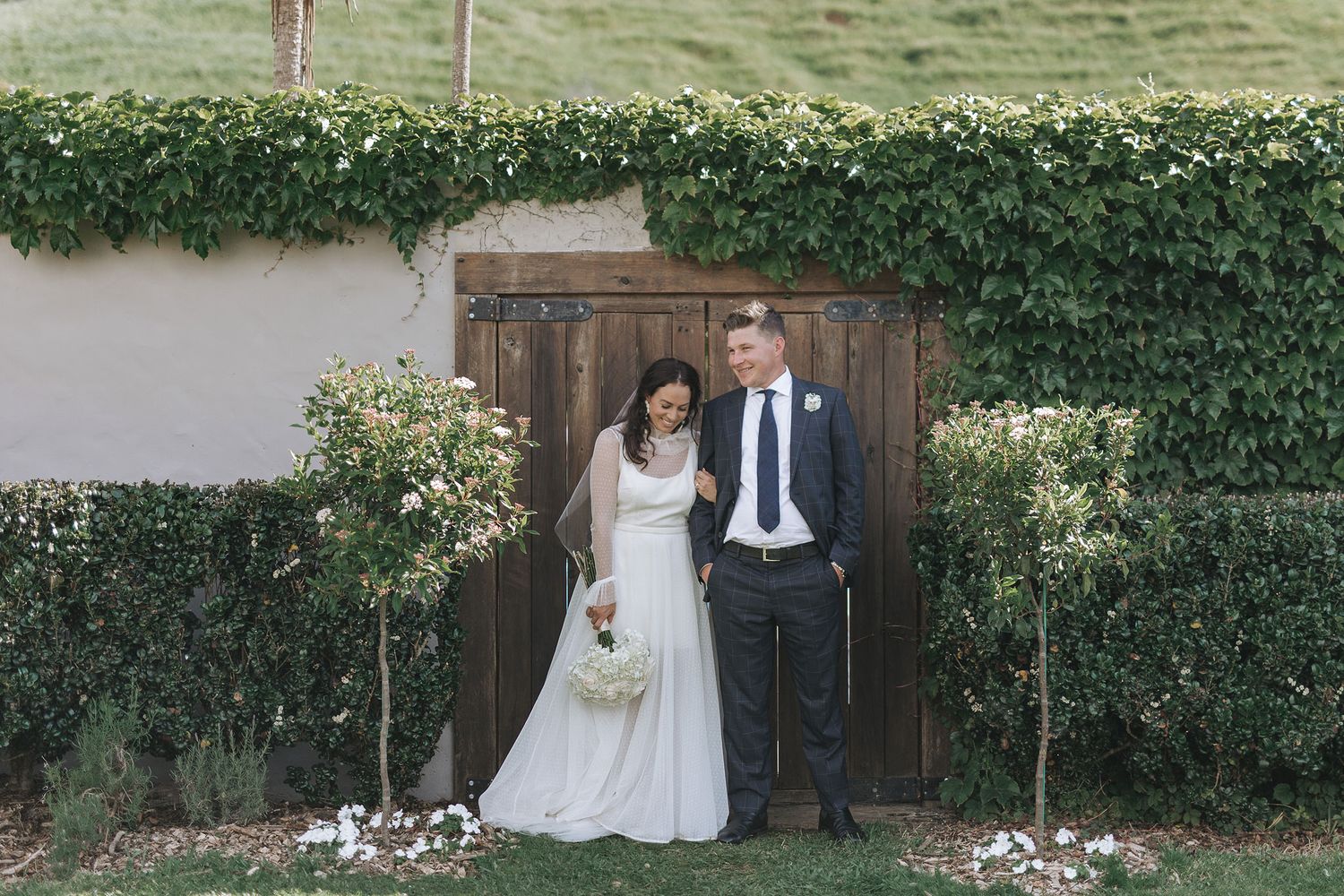 A wedding couple stands in front of a rustic wooden door surrounded by green ivy and white blooming flowers.