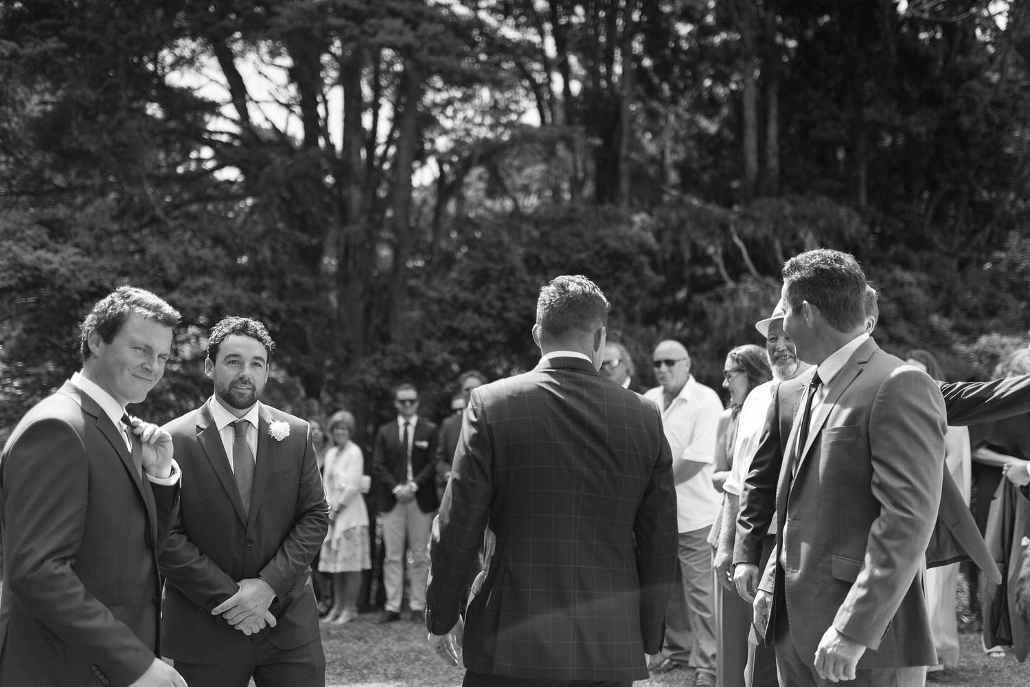 A group of wedding party guests gathered outdoors in formal attire laughing and socializing at a celebratory event.