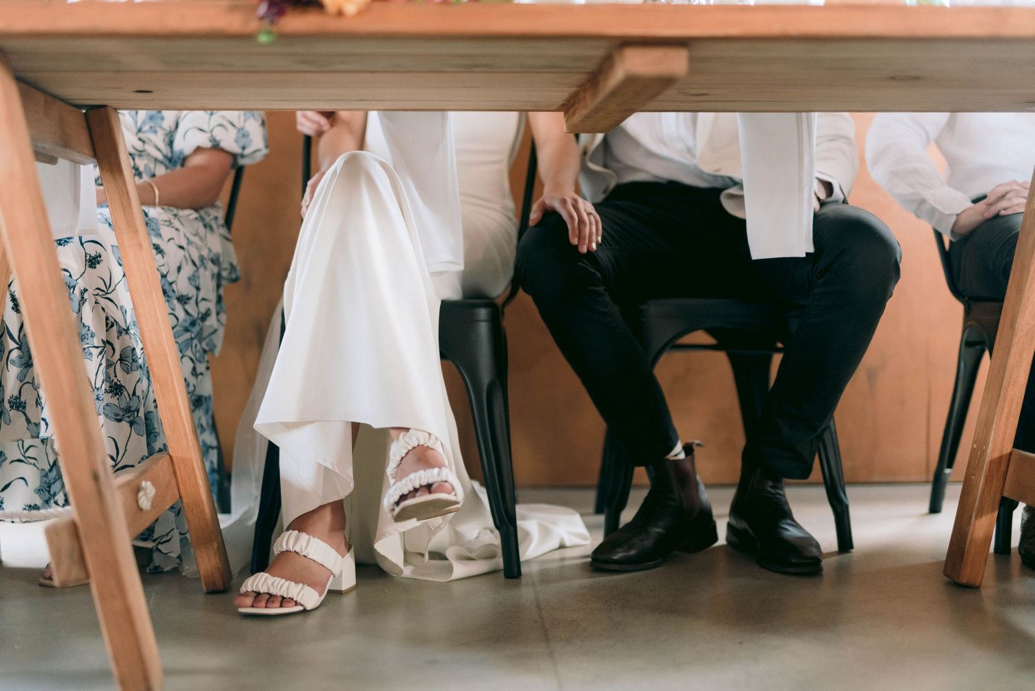 Wedding reception detail showing dress and formal attire under rustic wooden table.