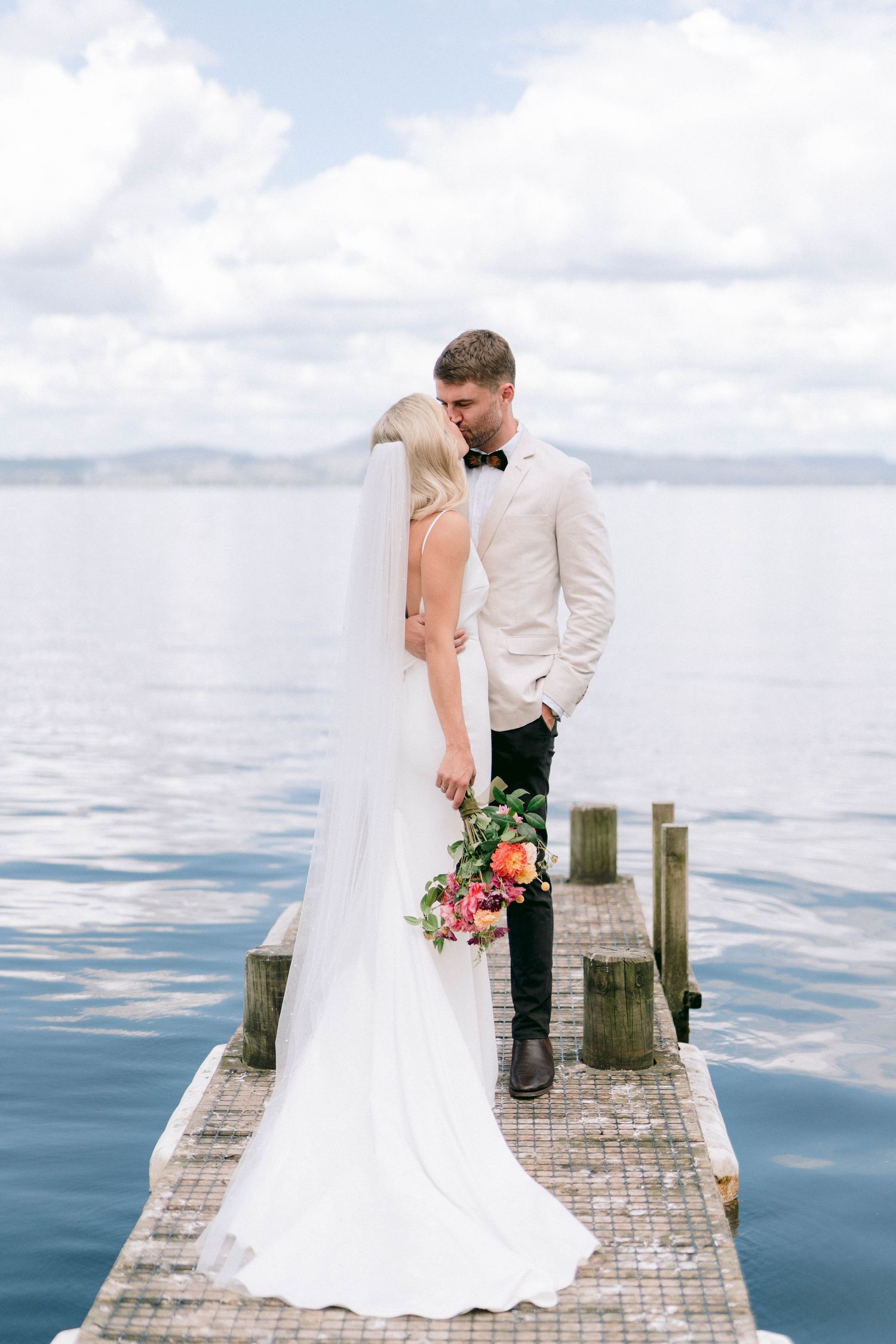 A beautiful wedding portrait on a wooden jetty with calm waters and cloudy skies in the background.