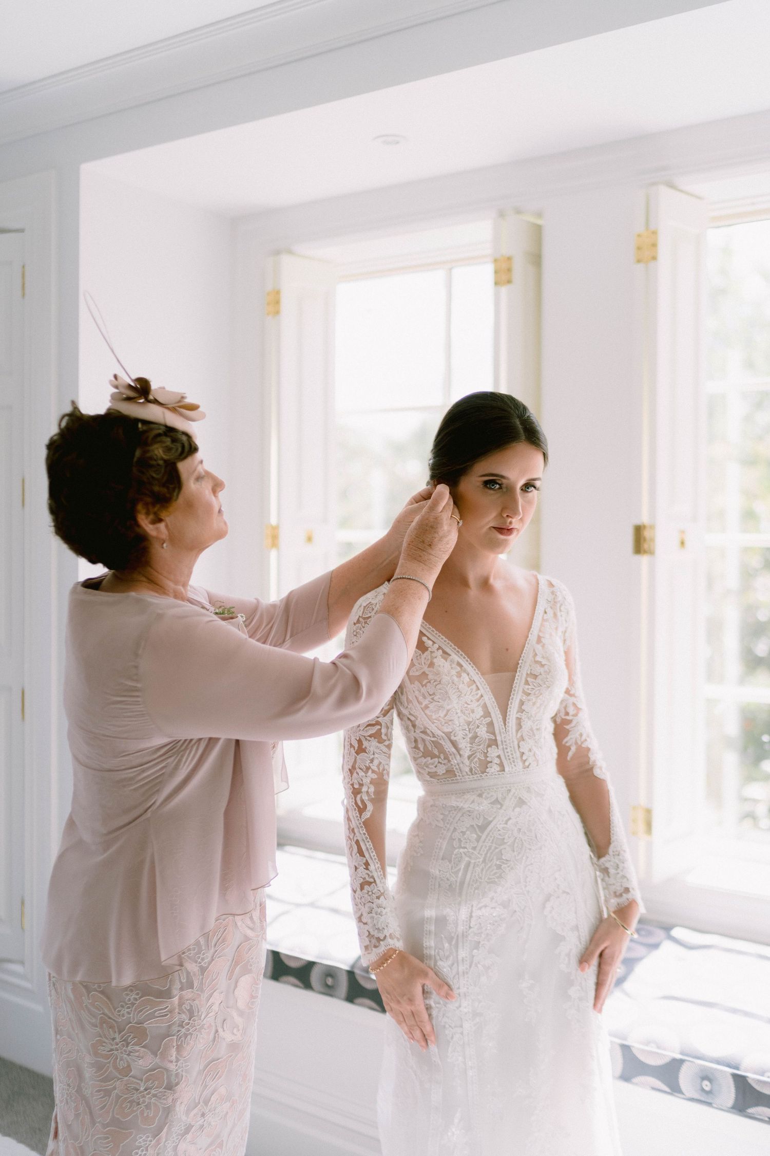 Bride getting dressed in a classic lace wedding gown while being helped in a bright sunlit room.