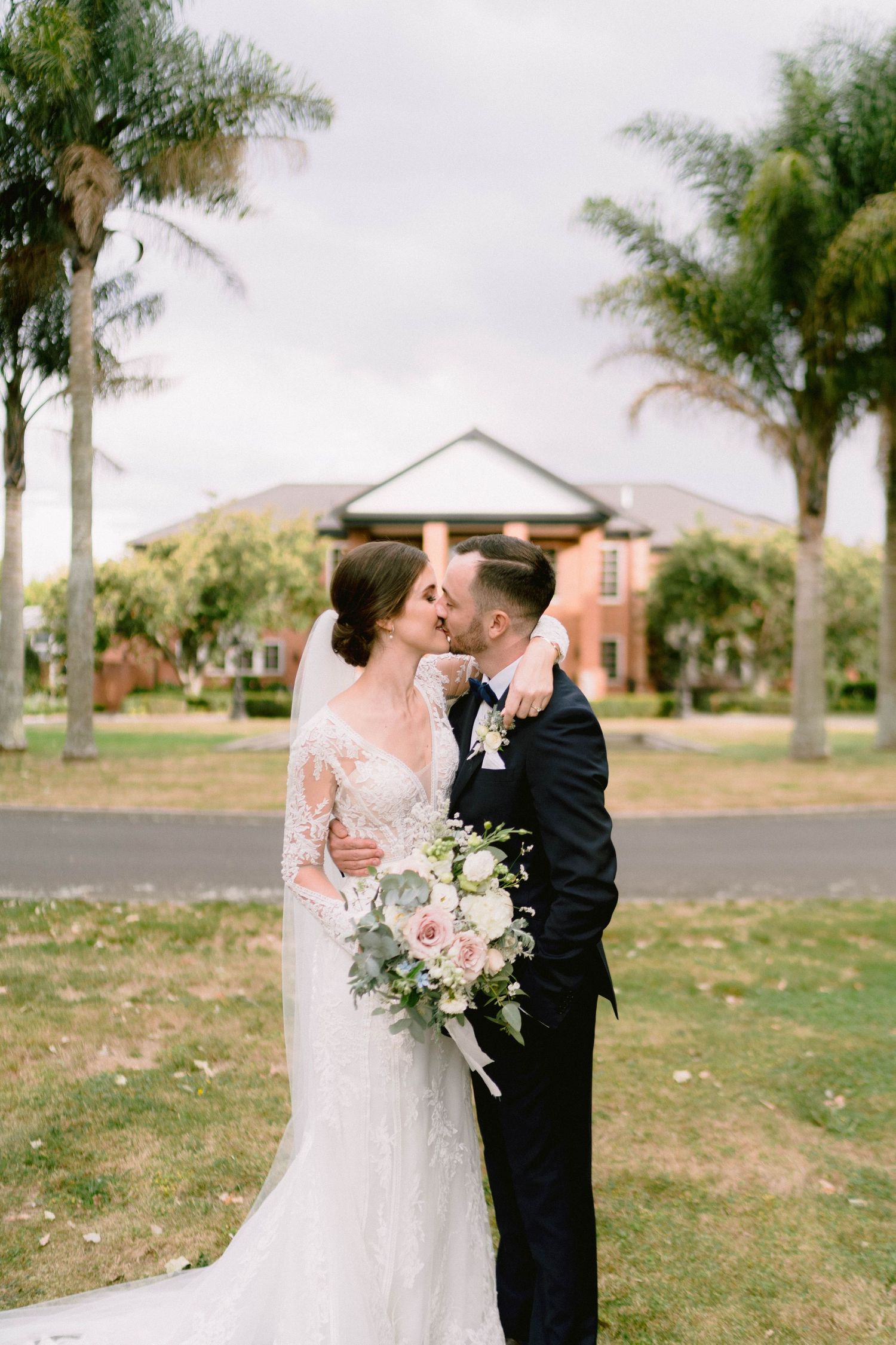 Newlyweds share a kiss with palm trees and buildings in background.