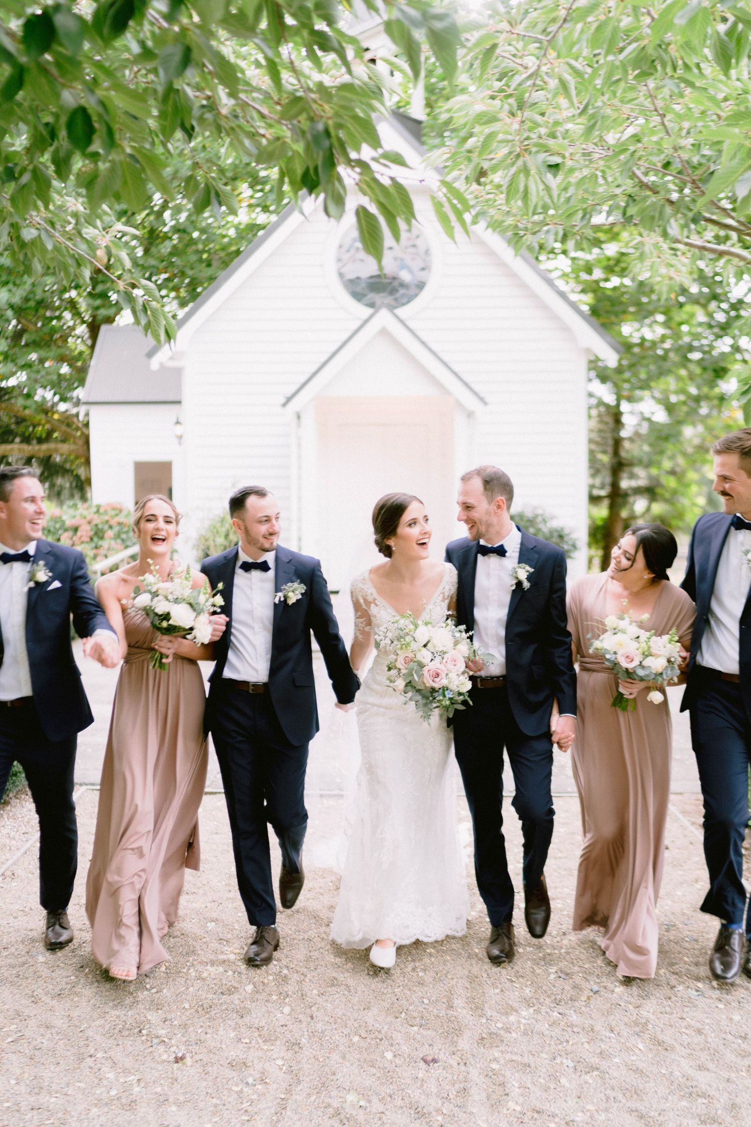 Wedding party in blush and black formal attire walking and laughing together outdoors.
