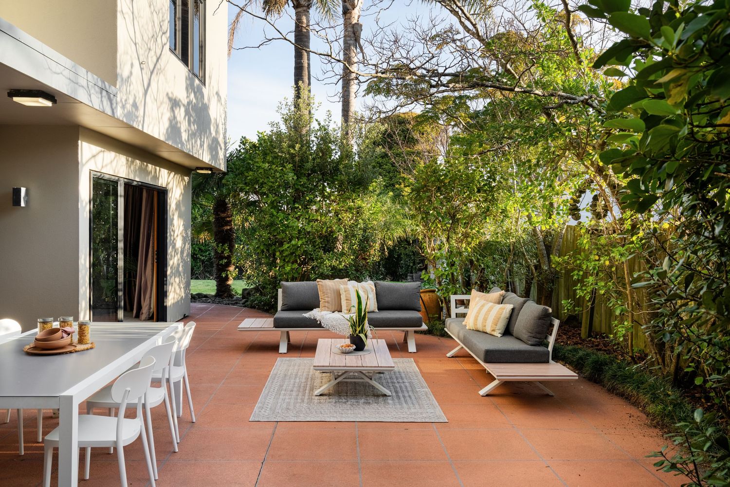 Outdoor patio with modern white furniture, gray cushions, coffee table, area rug, and lush green garden surroundings.