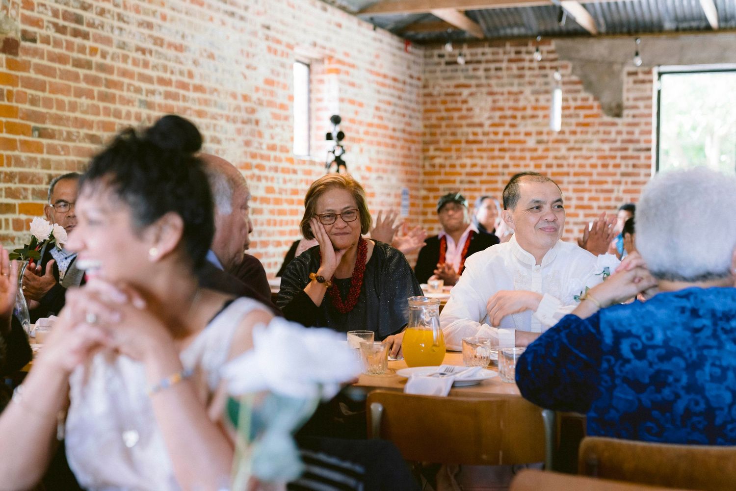 Wedding reception guests enjoying themselves at wooden tables inside a rustic brick-walled venue.