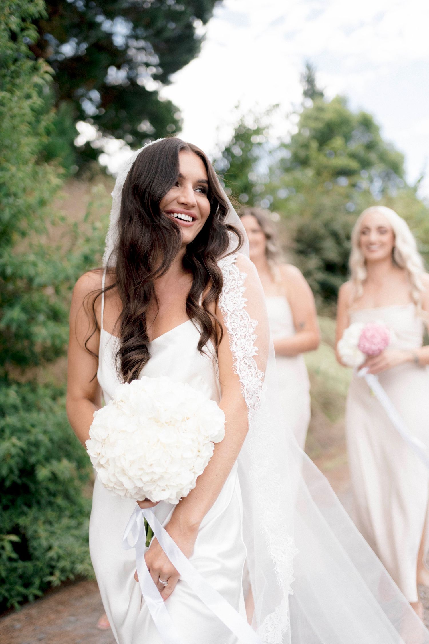 Bride in flowing white dress holds bouquet while walking outdoors with bridesmaids.
