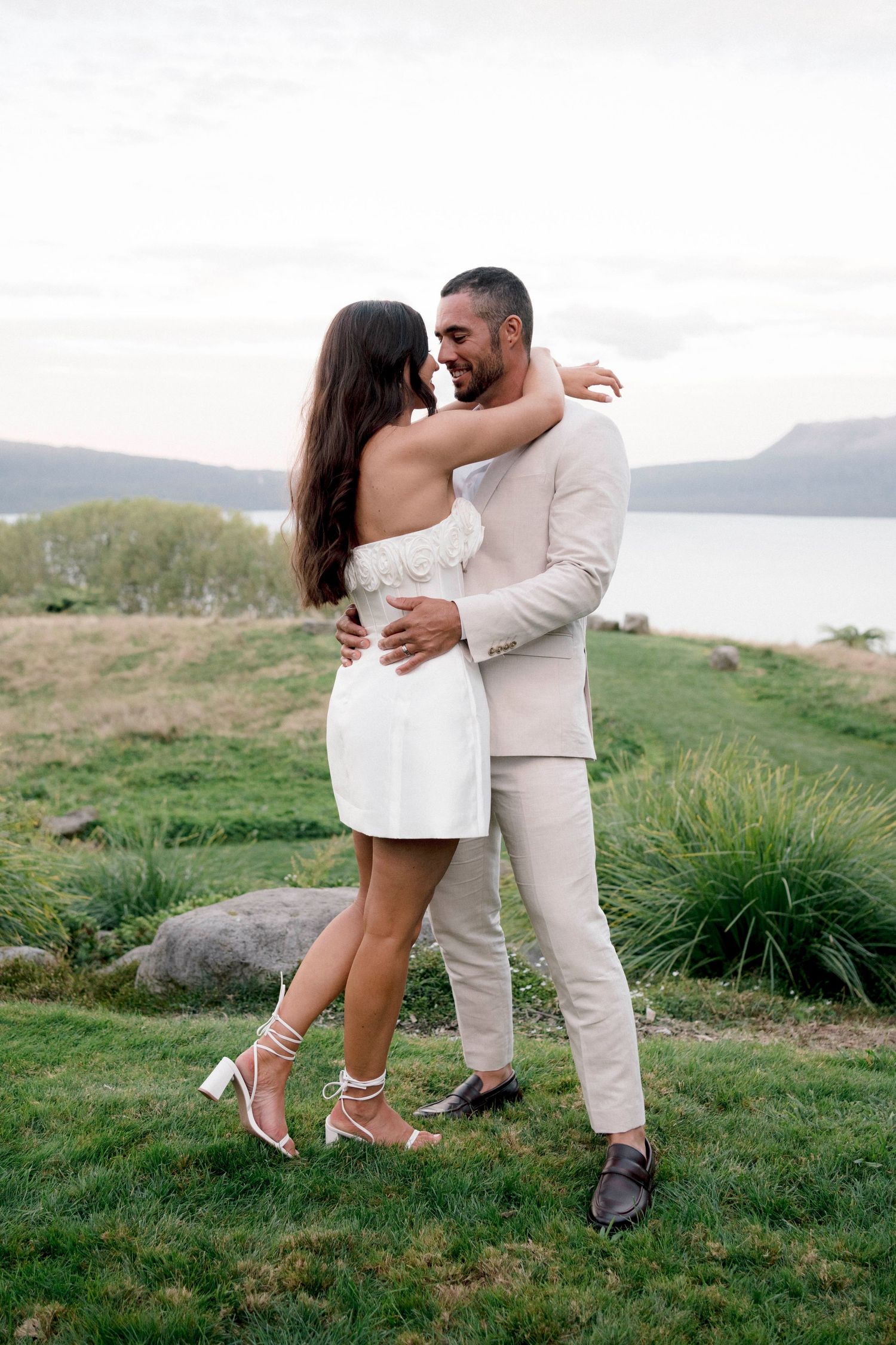 A romantic couple embraces on a grassy hilltop overlooking a lake, dressed in coordinating cream-colored outfits.