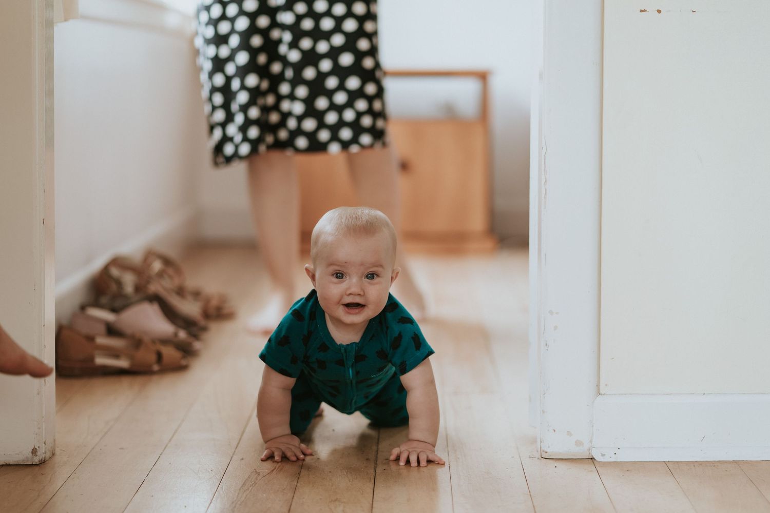 Small baby in polka dot outfit crawls in bright hallway while exploring the wooden floor.