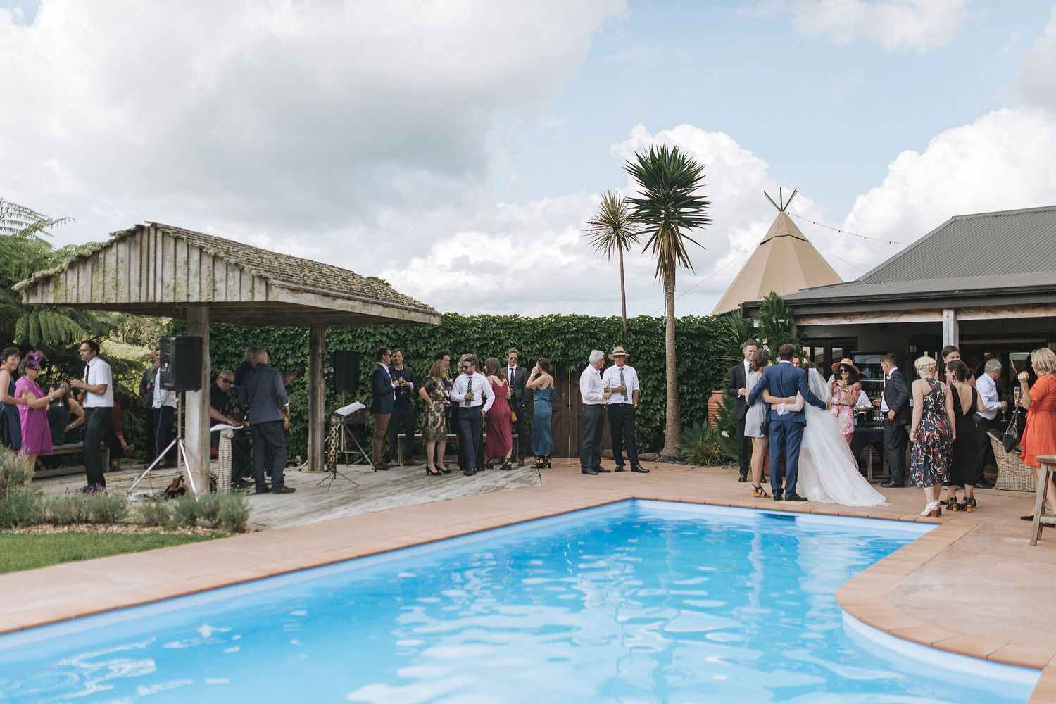 Wedding guests gather around an outdoor swimming pool at a venue with tropical landscaping and buildings on a sunny day.