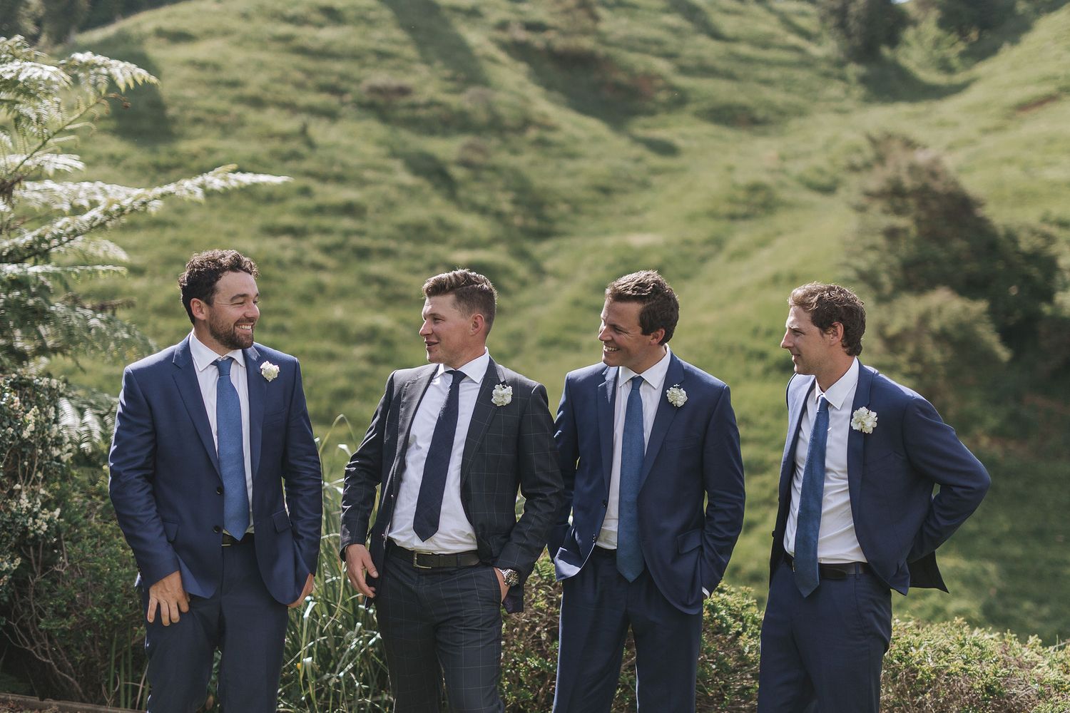 A group of groomsmen in matching navy suits walk and laugh together on a scenic grassy hillside for wedding photos.
