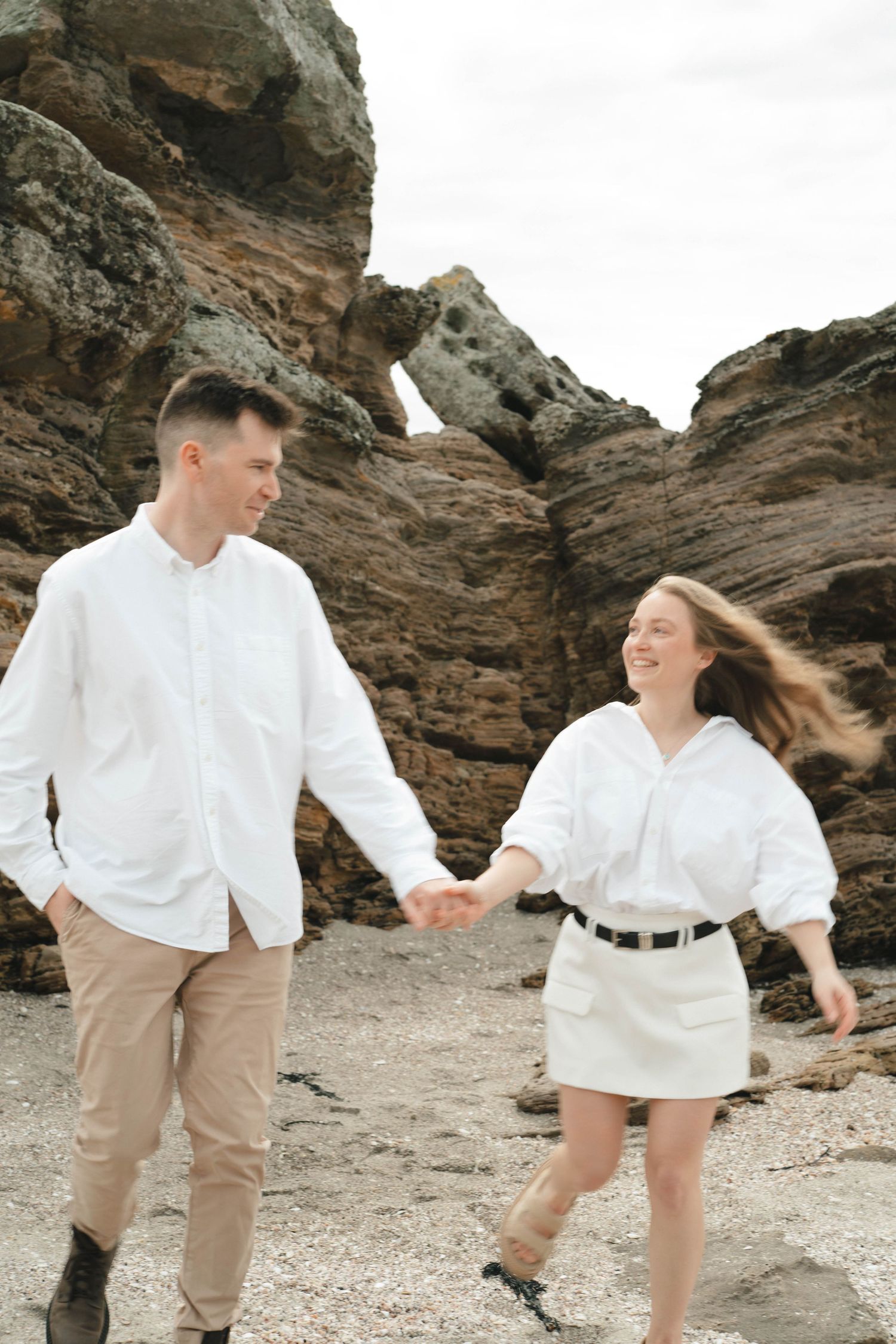 Couple in white outfits dancing and spinning together on rocky beach.
