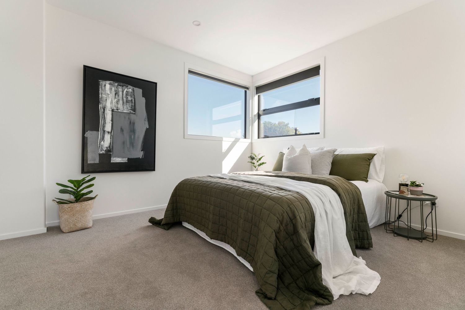 Modern bedroom with corner windows, olive green bedding, grey carpet and black and white artwork on white walls.