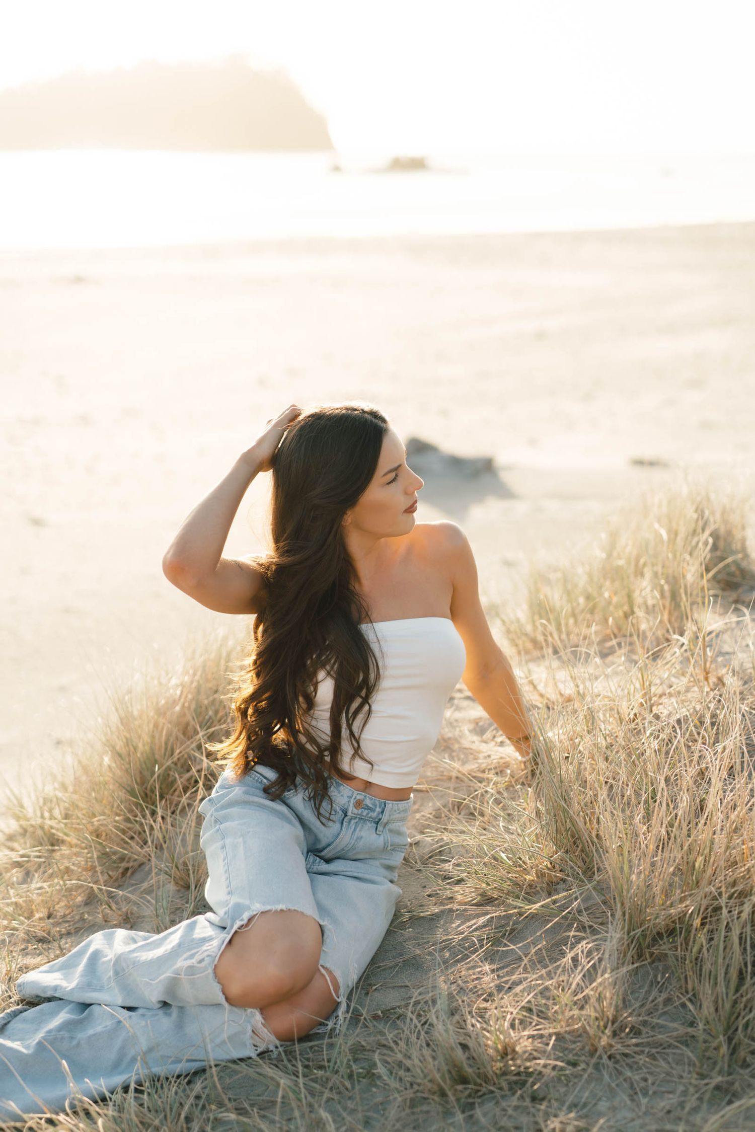 Woman in white top and denim relaxing in tall grass during golden hour at sunset.