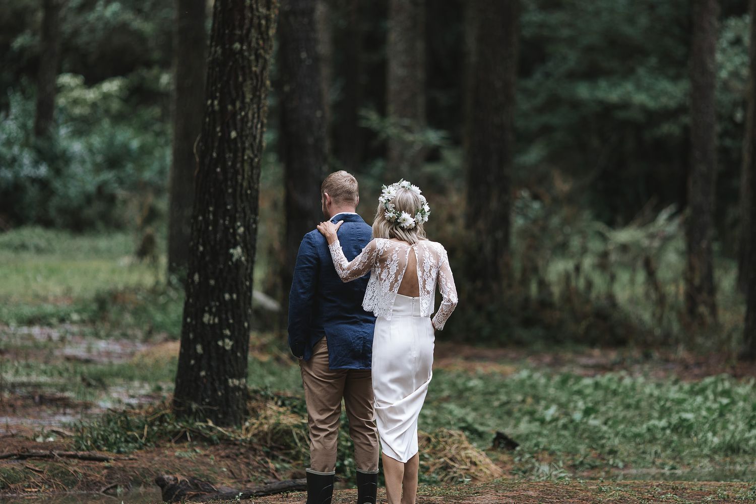 A couple walks together between tall trees on a forest path wearing wedding attire.