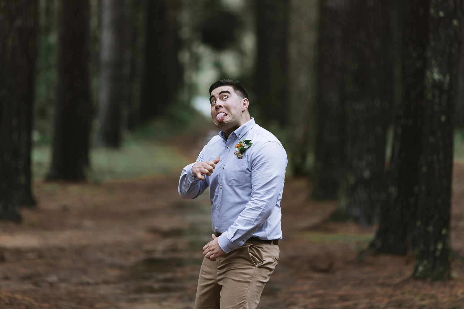 A groom wearing a light gray suit poses playfully near forest trees at an outdoor wedding photoshoot.