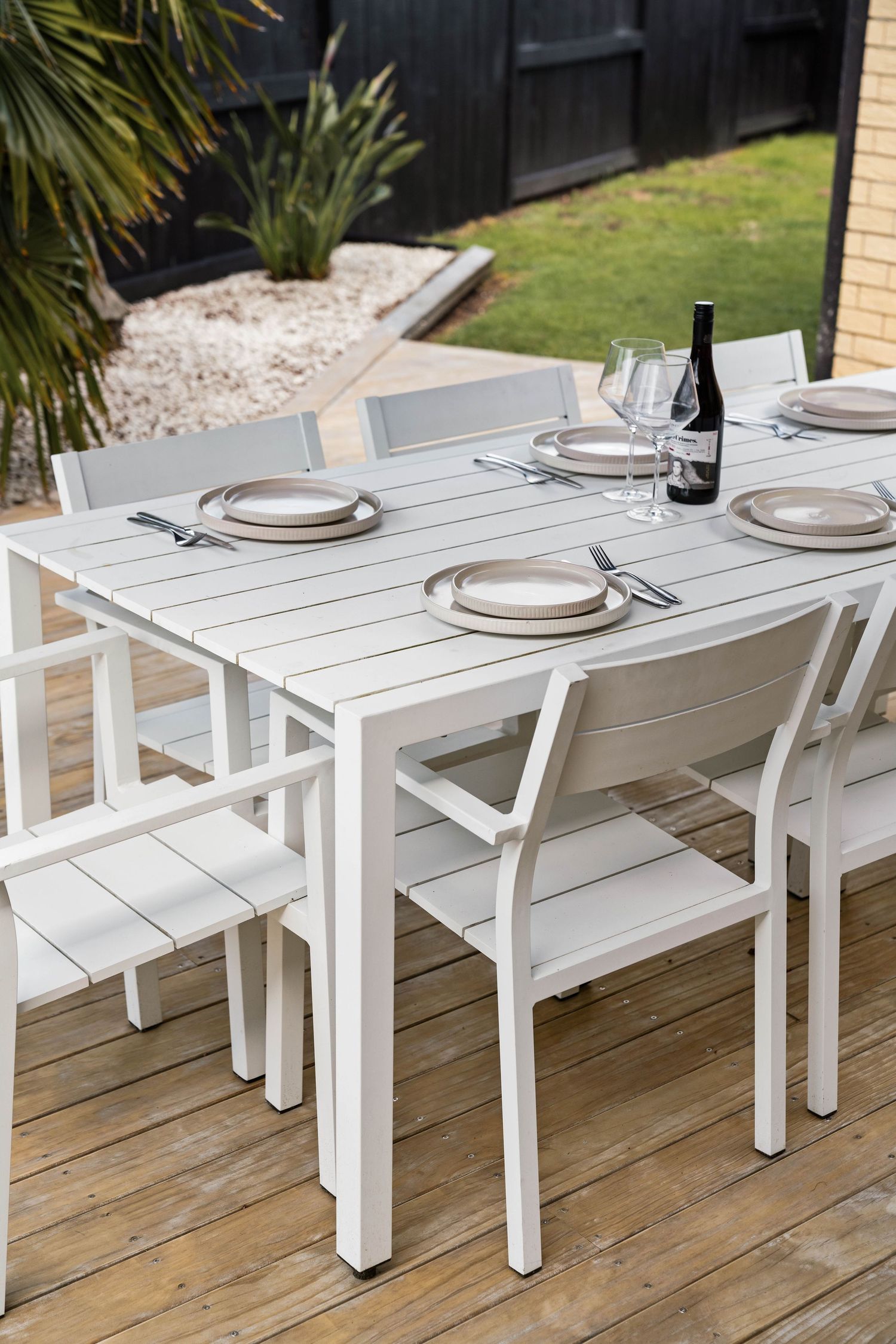 White outdoor dining set with place settings and wine bottle on wooden deck with tropical garden view.
