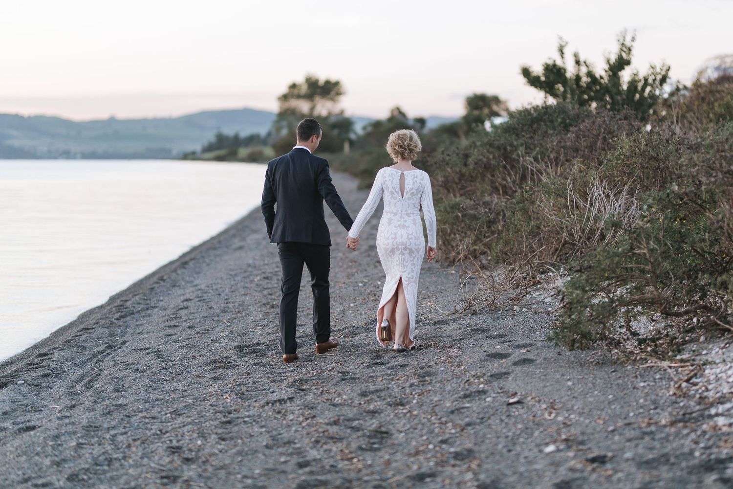 A couple walks hand in hand along a rocky beach path at sunset with water and mountains in the background.