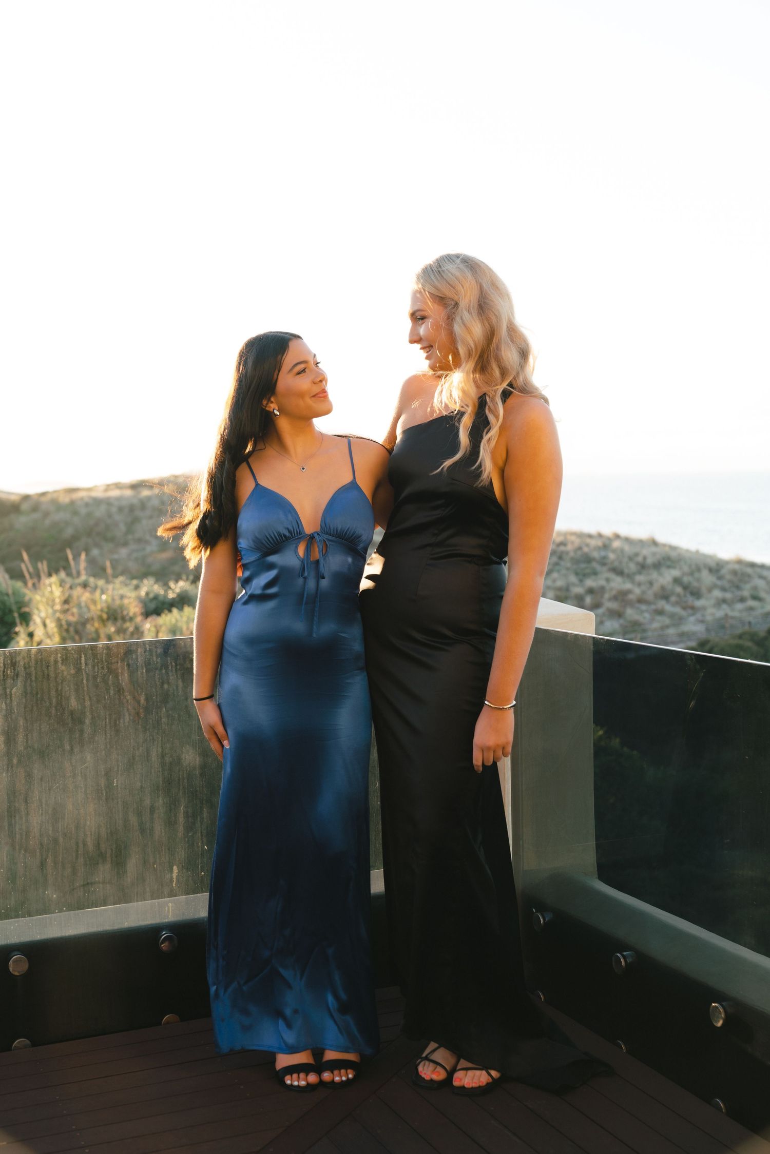 Two friends in evening dresses sharing a moment on outdoor terrace at sunset.