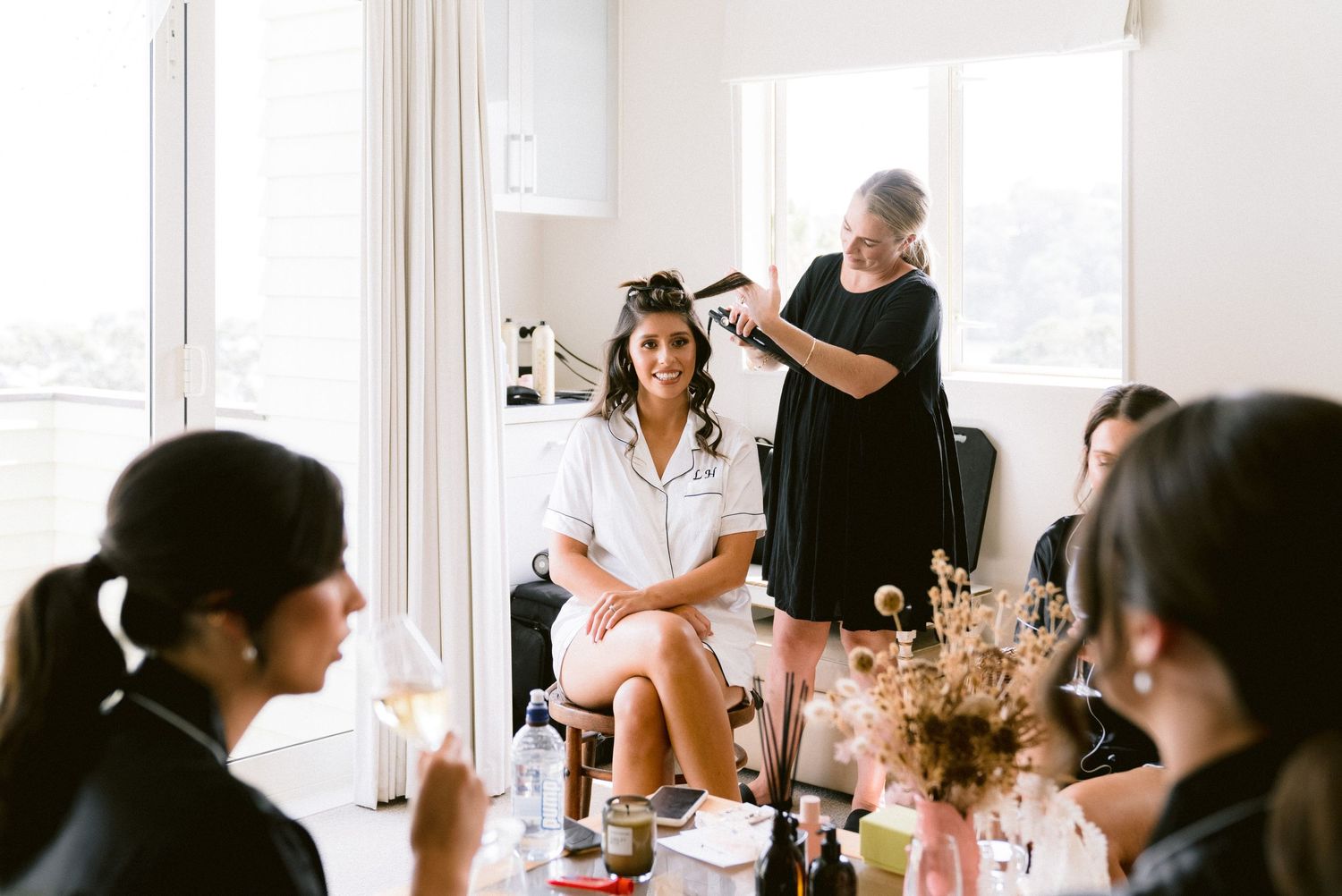 Stylist works on client's hair while others gather around a decorative table for bridal preparation before wedding ceremony.