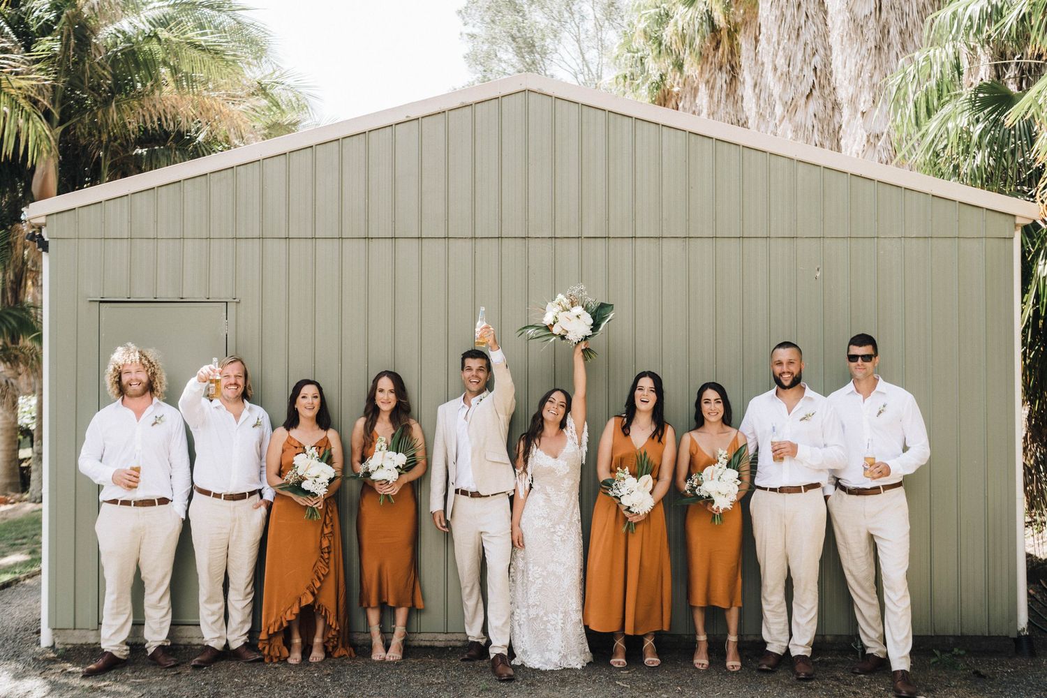 Wedding party poses together in front of a gray barn building wearing rustic style attire.
