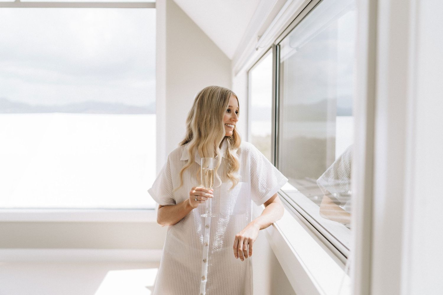 Woman in white dress standing by bright window with sheer curtains in modern minimalist room.