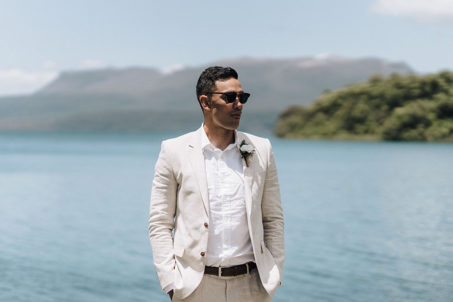 A person in a light colored suit stands against a backdrop of mountains and lake waters.