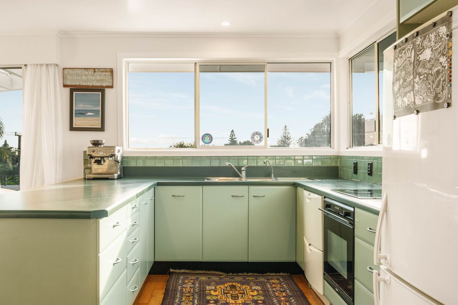 Modern L-shaped kitchen with mint green cabinets, white countertops, and large windows providing natural light.