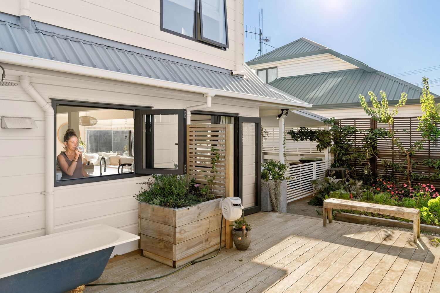 Modern white house exterior with wooden deck, planter boxes, and garden featuring a large picture window and metal roof.