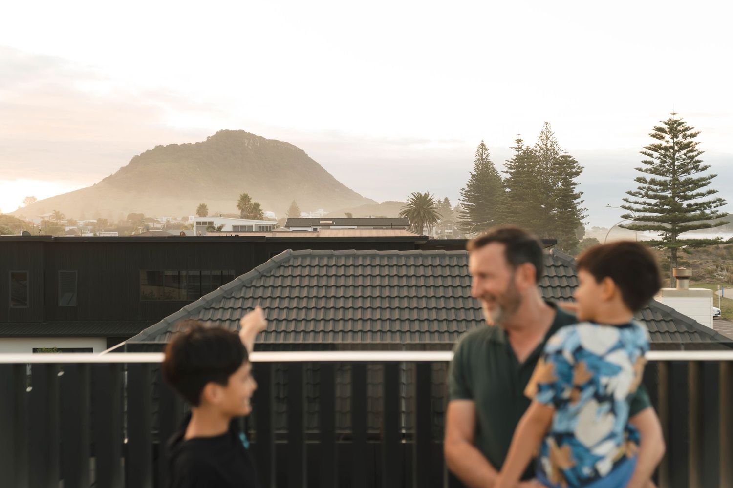 A group stands on an outdoor deck with Mount Maunganui visible in the distance during sunset.