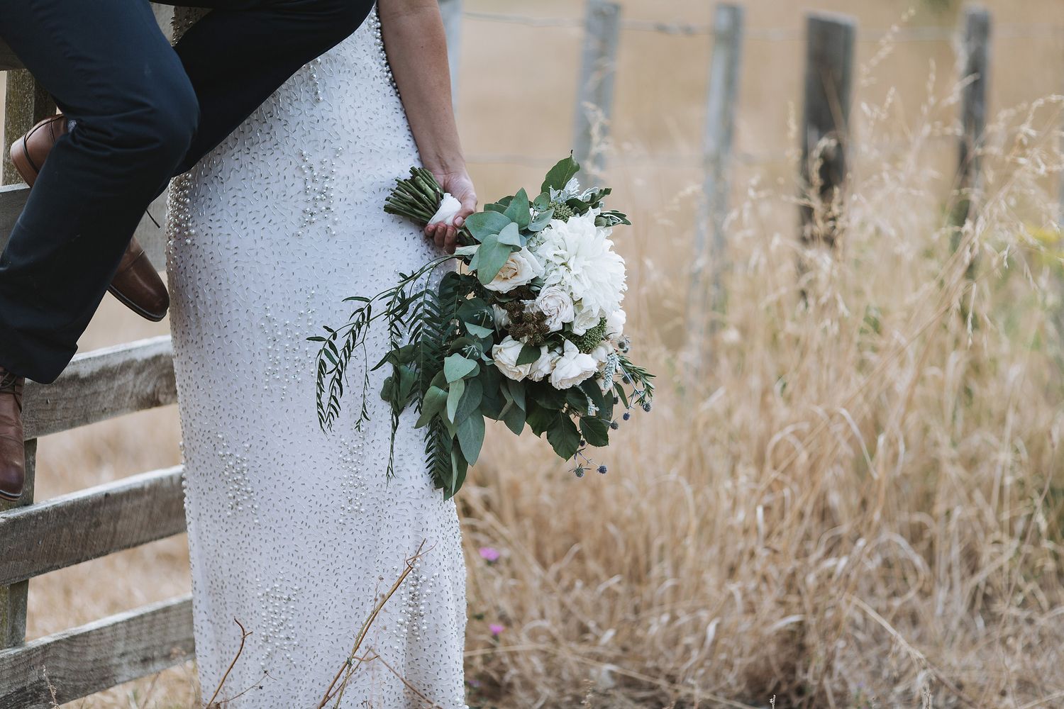 A couple holds hands while walking through a sunlit field of tall dried grass wearing elegant white wedding attire.