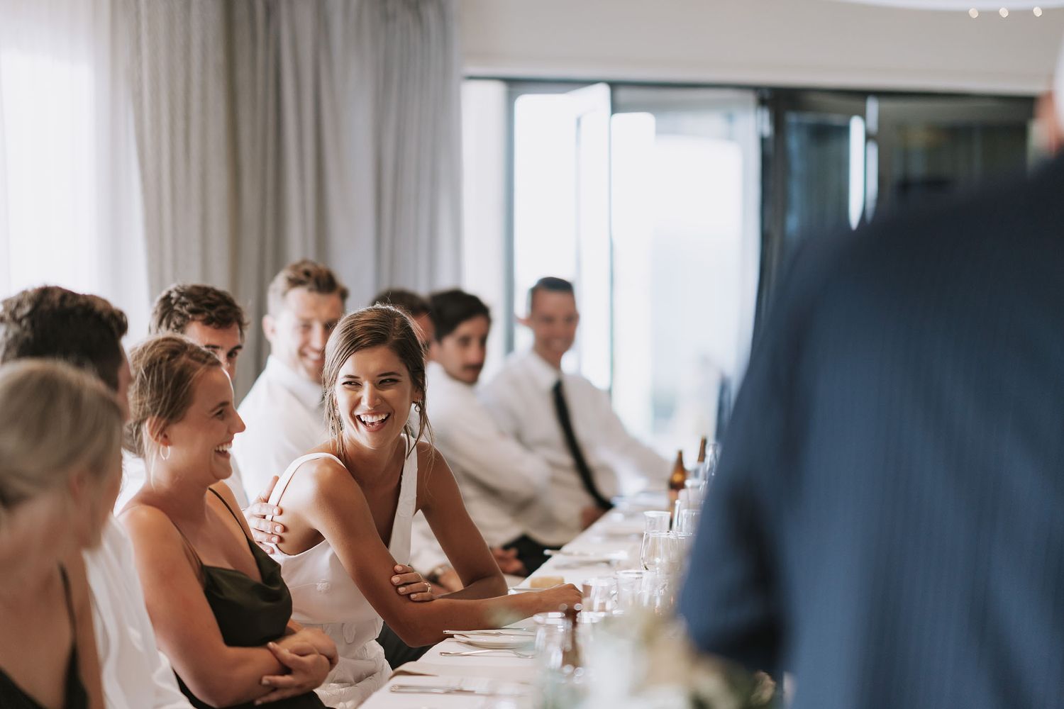 Wedding party members sit together at a long table during a reception, smiling and enjoying drinks.