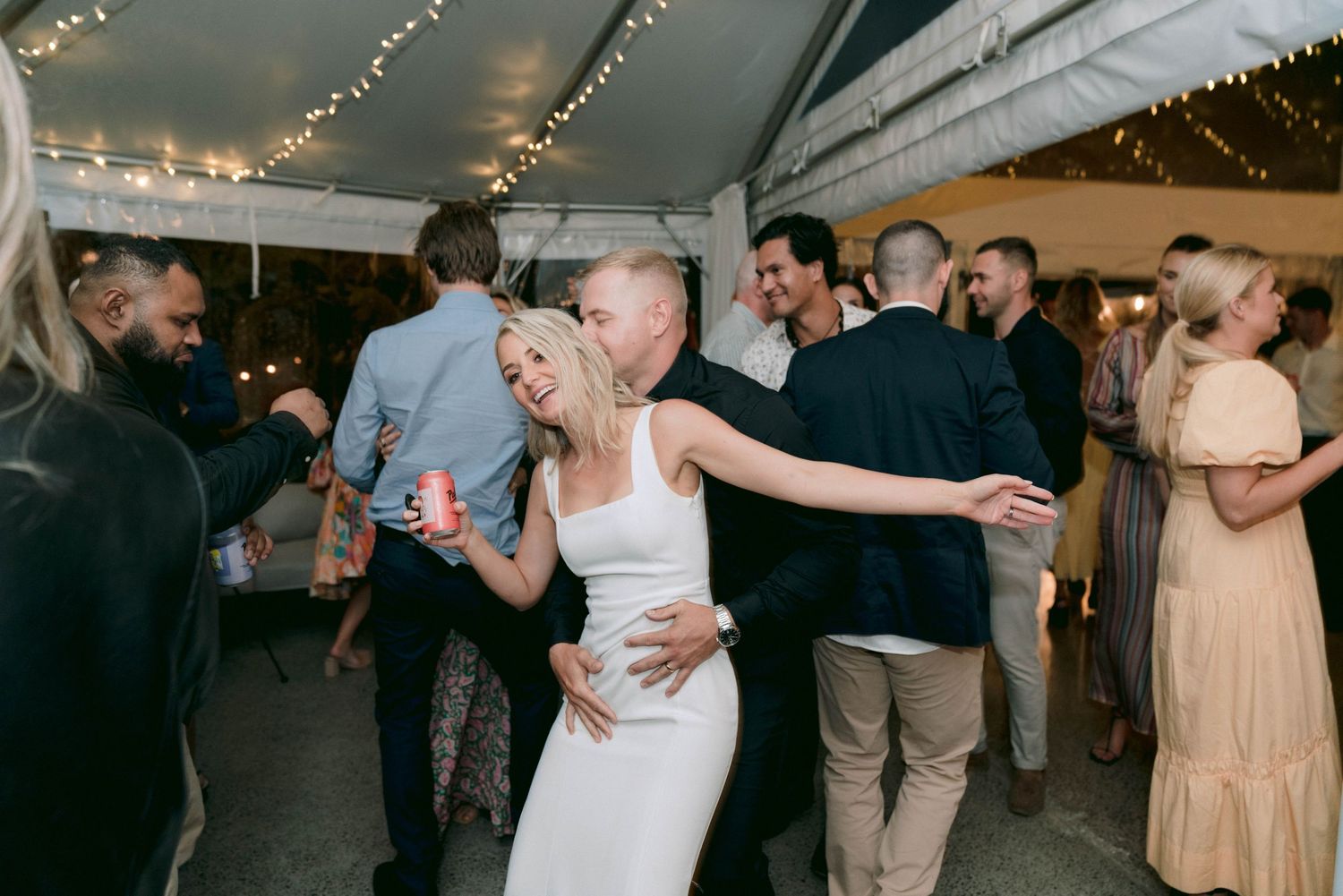 Party guests celebrating and dancing under a tent at an evening celebration with string lights overhead.