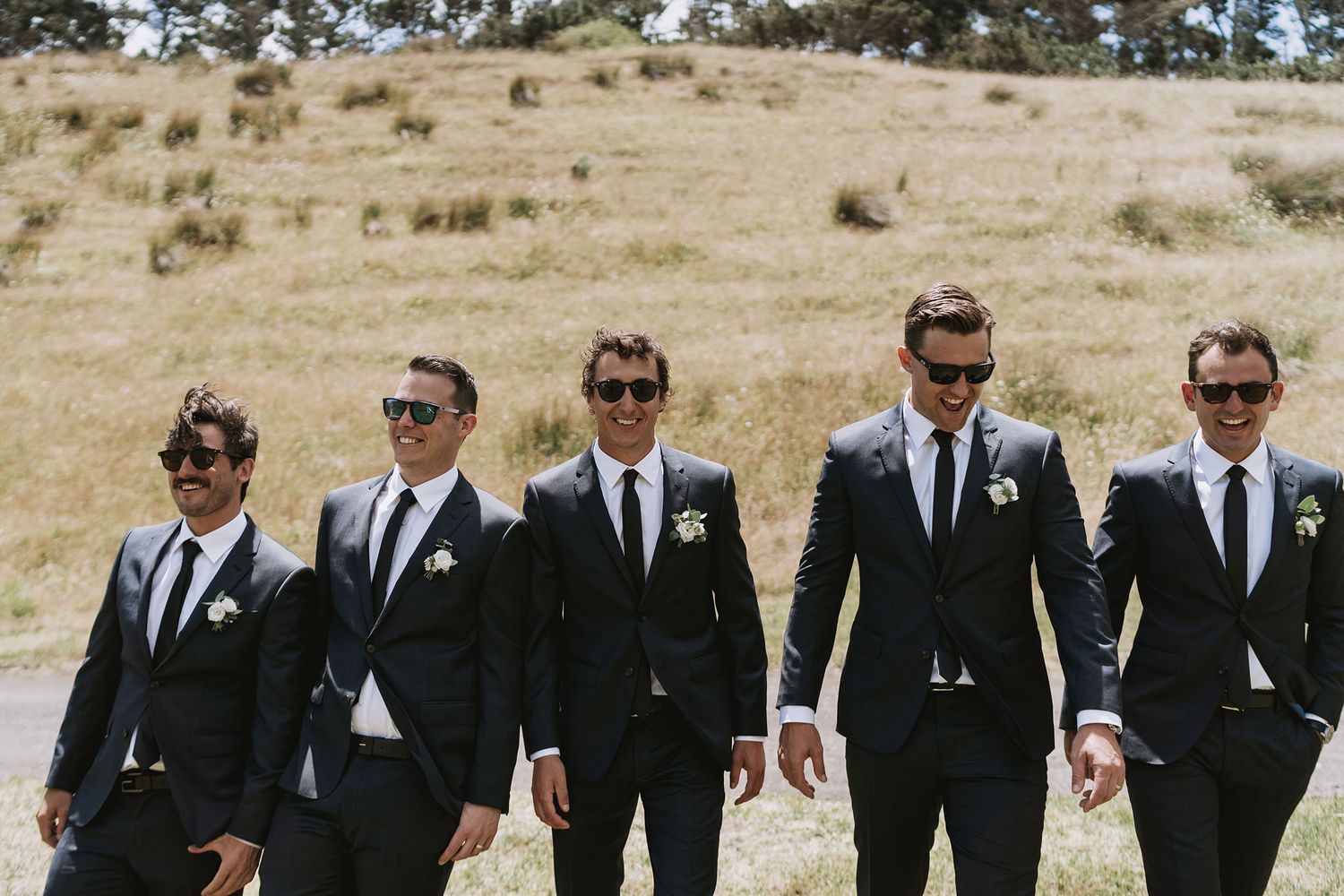 Groomsmen in black suits standing together on a grassy hillside, showing progression from serious to candid poses.