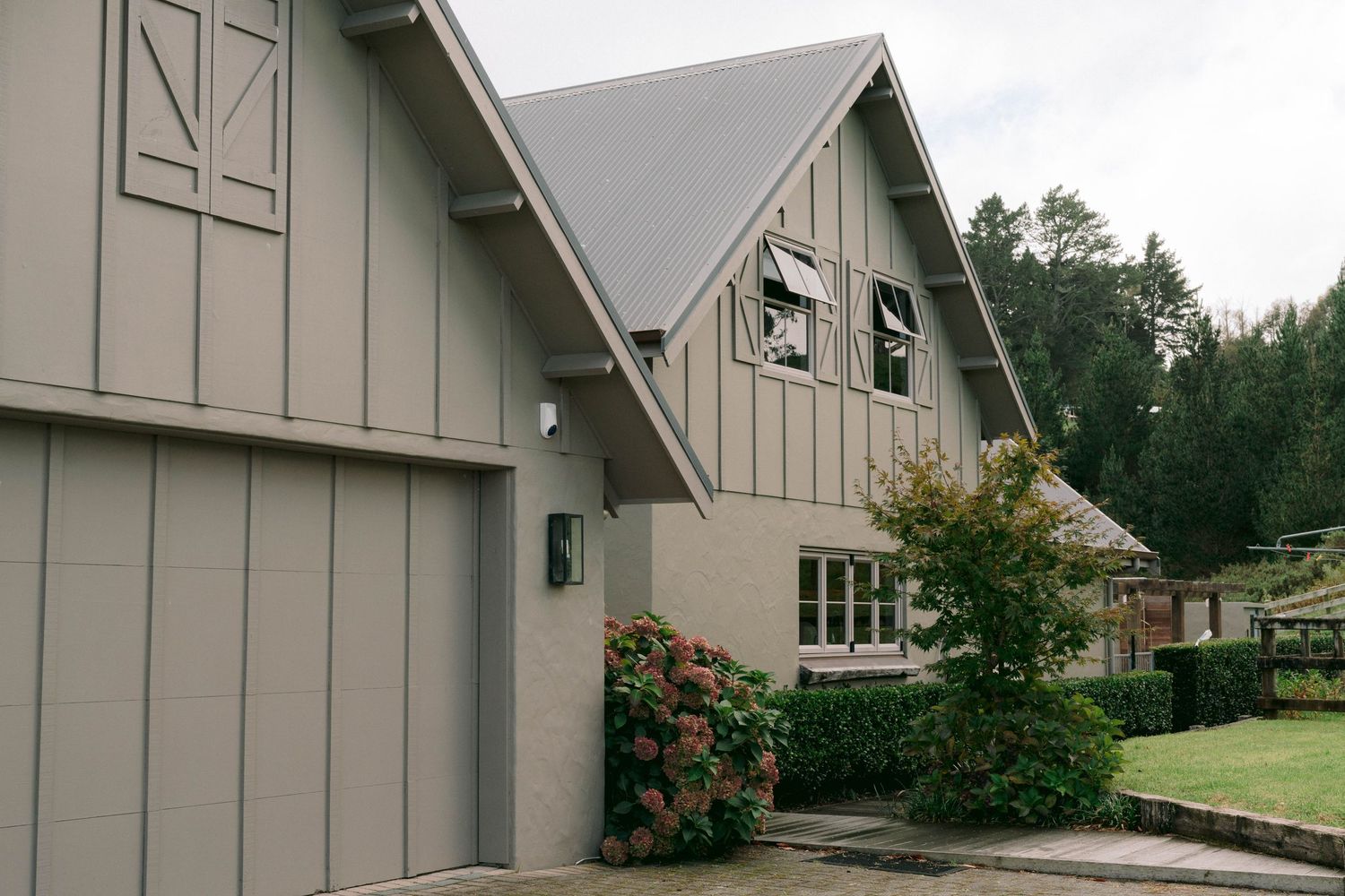 A beige contemporary house with steep gabled roofs, flowering bushes and evergreen trees in the background.