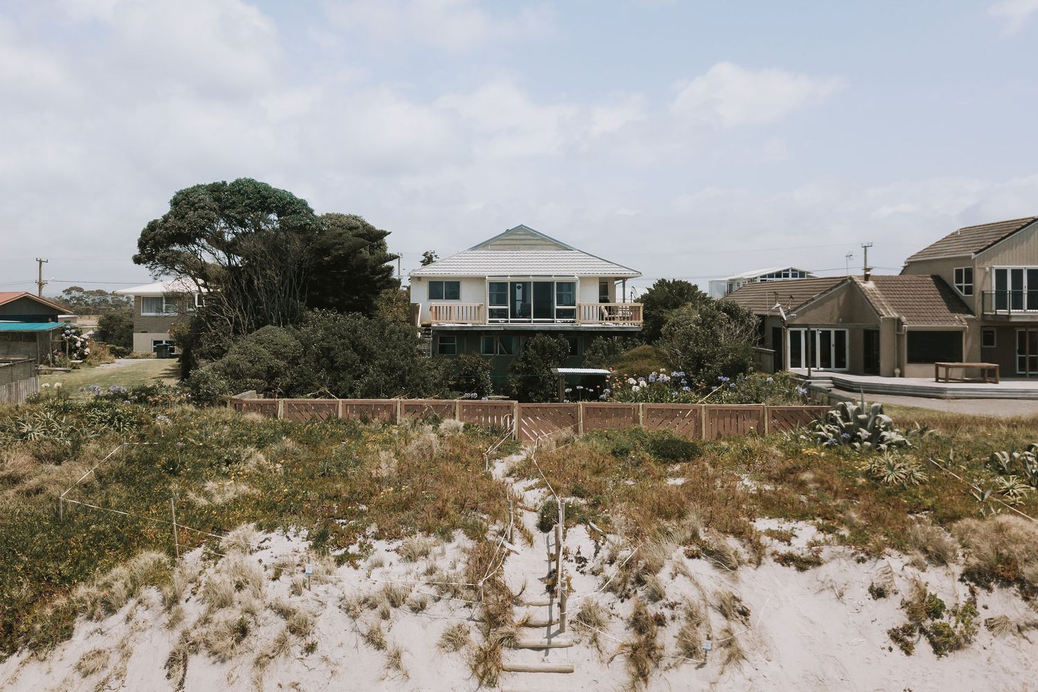 Coastal beach house with white siding sits elevated above sandy dunes on a sunny day.