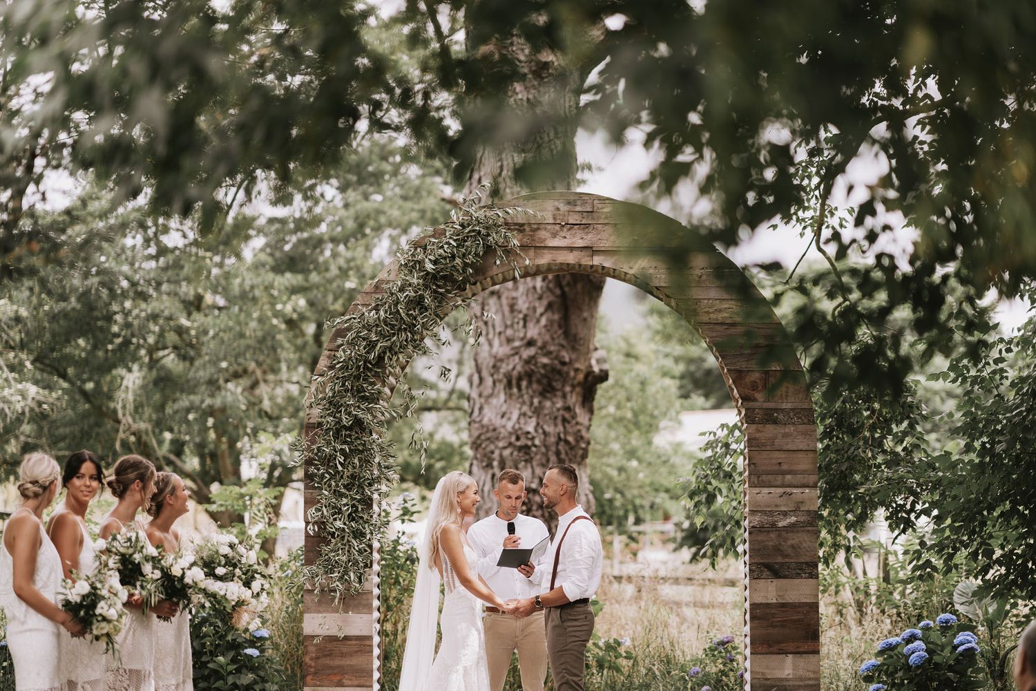 A couple embraces under oak trees in a dreamy outdoor garden wedding ceremony setting.