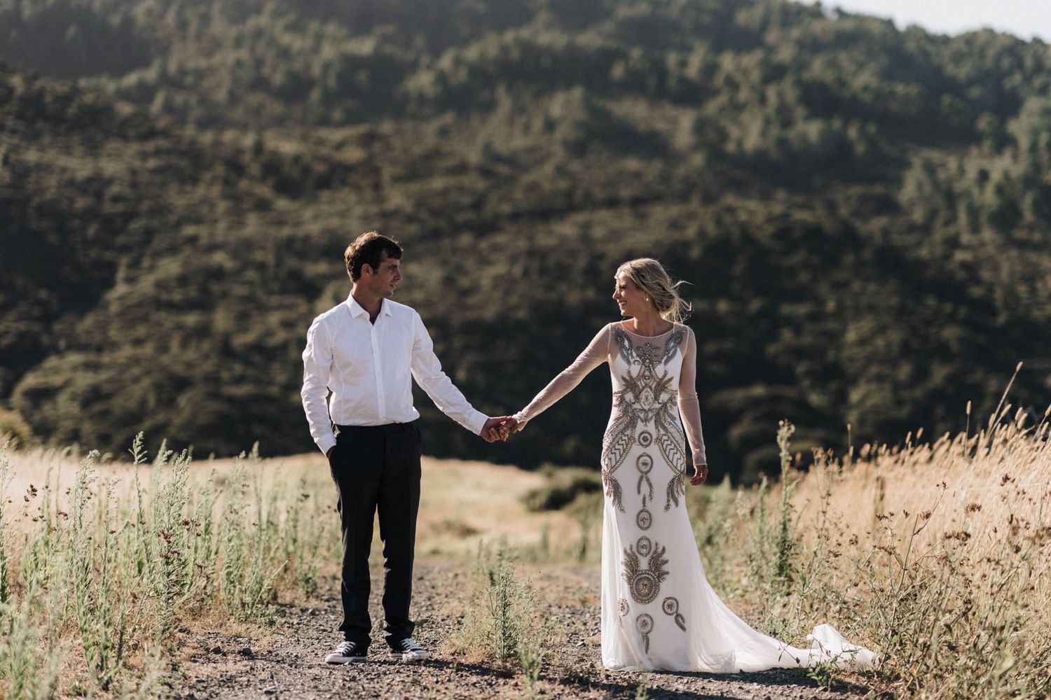 A couple in wedding attire walks together holding hands on a path through golden grass with mountains in background.