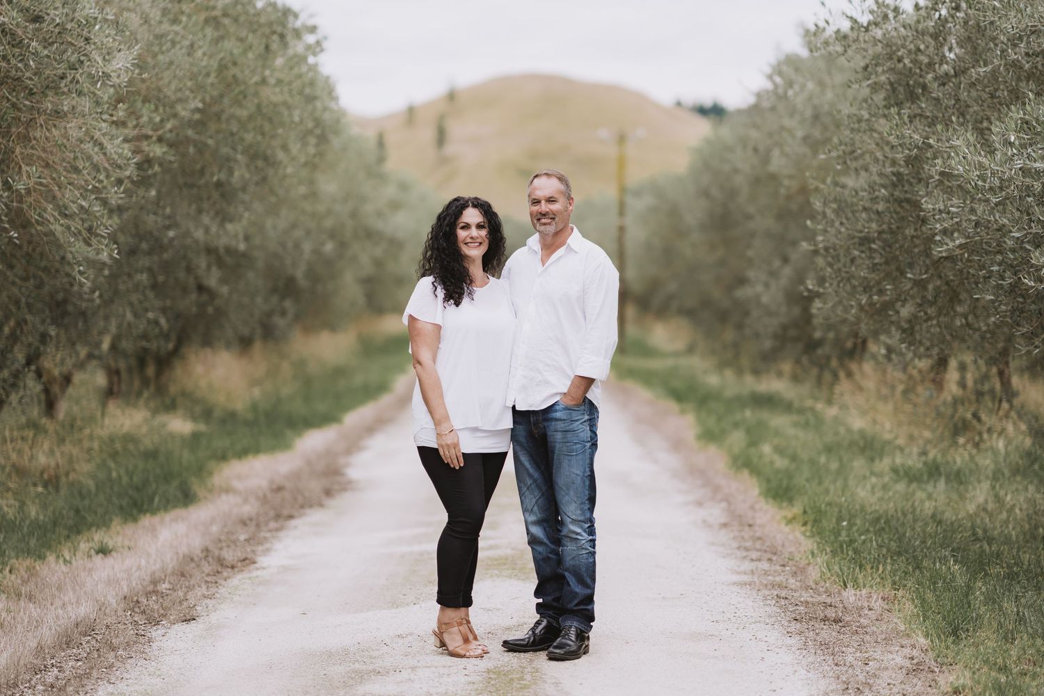 A couple stands together on a dirt path lined with olive trees in a rustic outdoor setting.