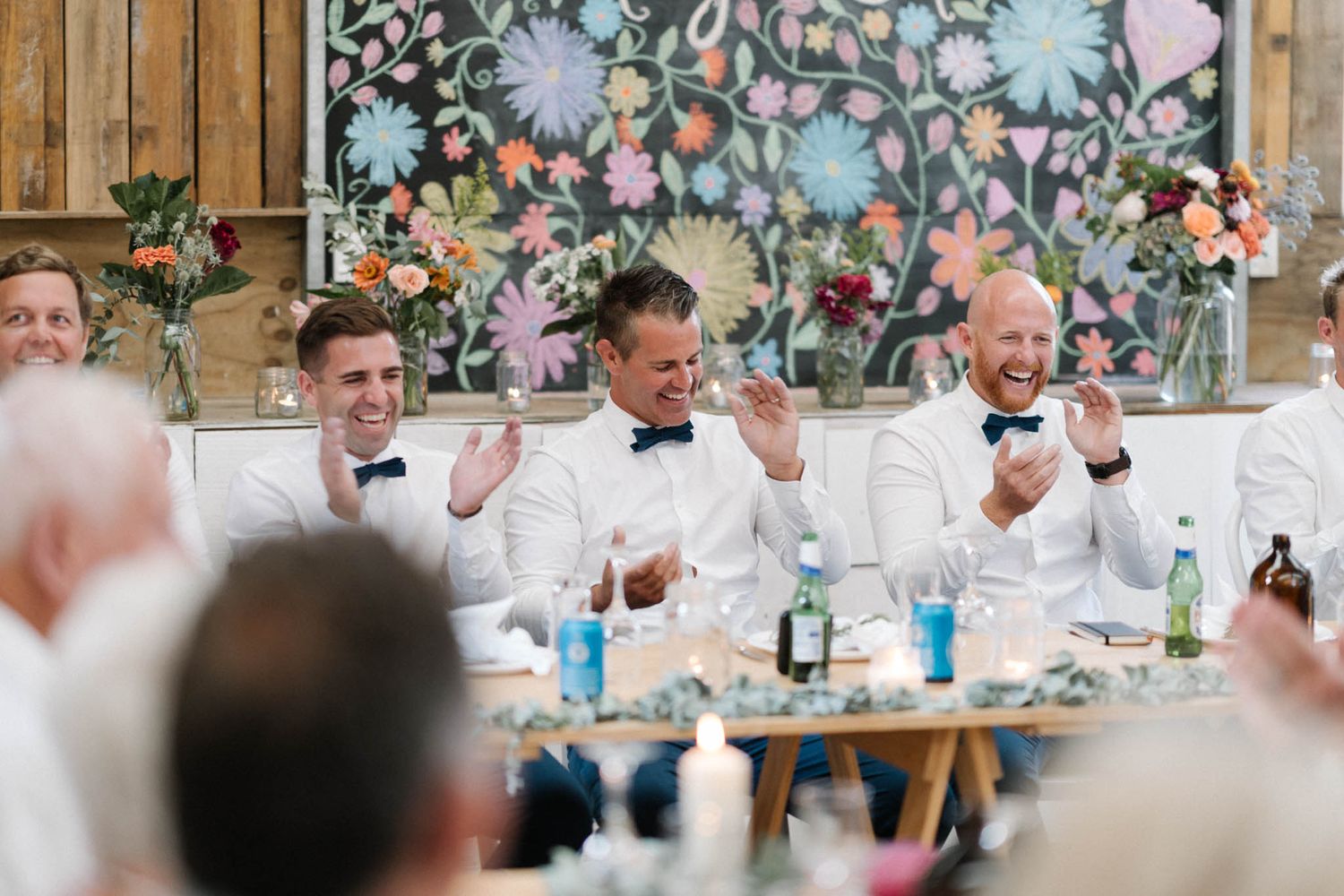 Wedding reception guests seated at long tables enjoy a festive meal and conversation in an elegant floral-decorated venue.