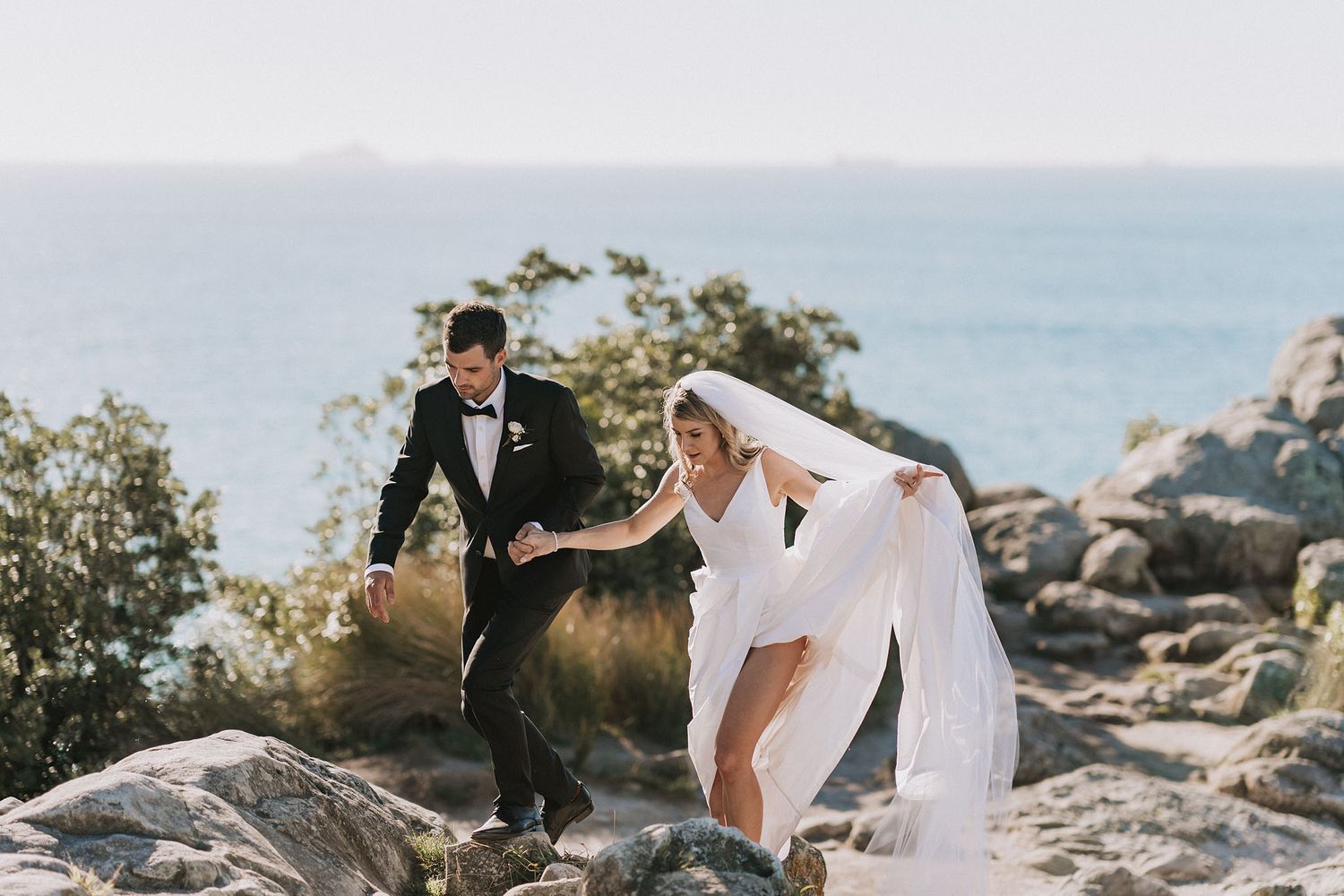 A newly married couple carefully walking on rocky coastal terrain with ocean views and sunlit foliage in the background.