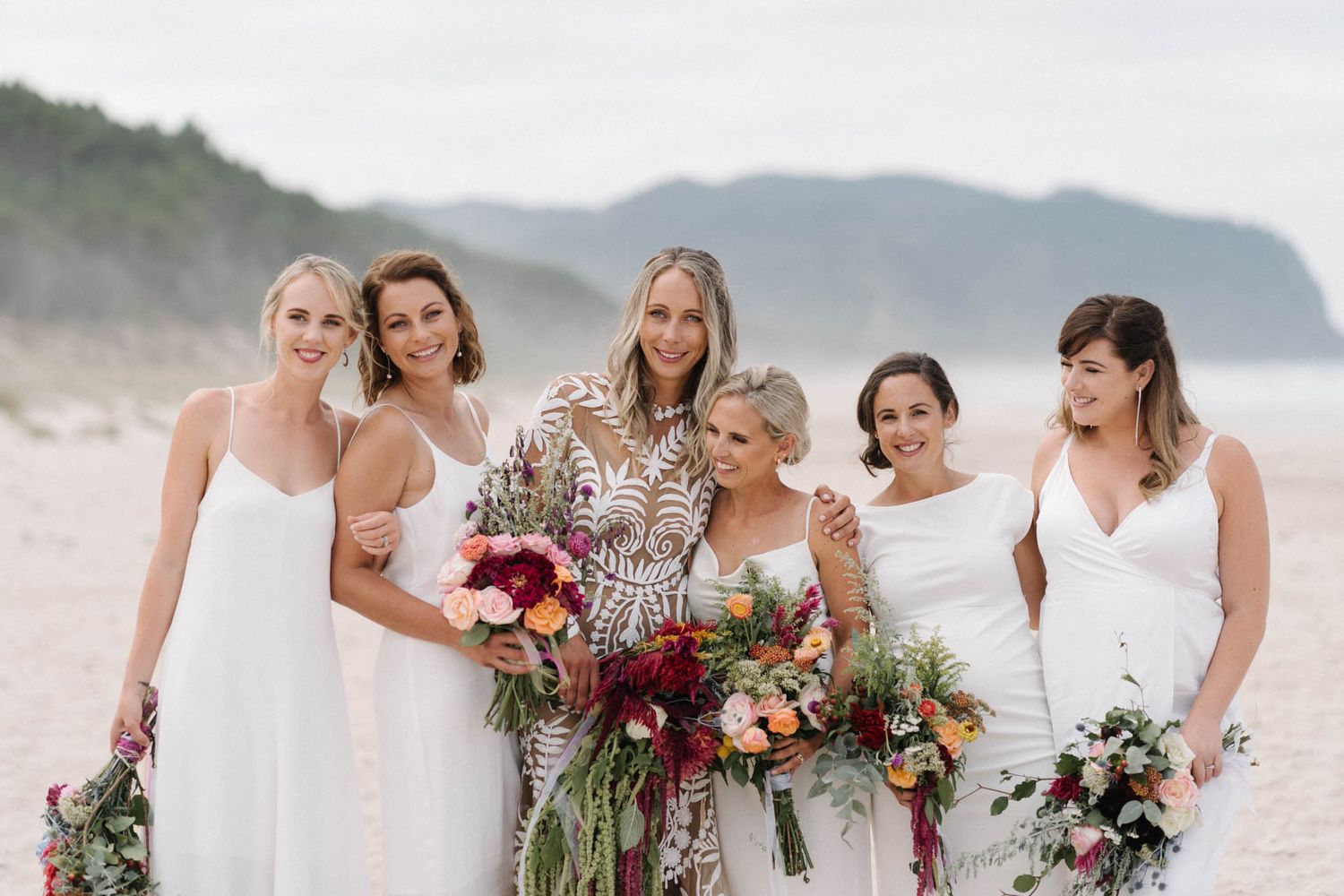 Beautiful bridesmaids in white dresses hold vibrant burgundy and blush bouquets on a misty beach with mountains.
