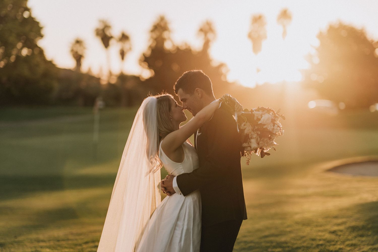 A couple in wedding attire embrace in a golden sunlit garden during sunset with palm trees in the background.