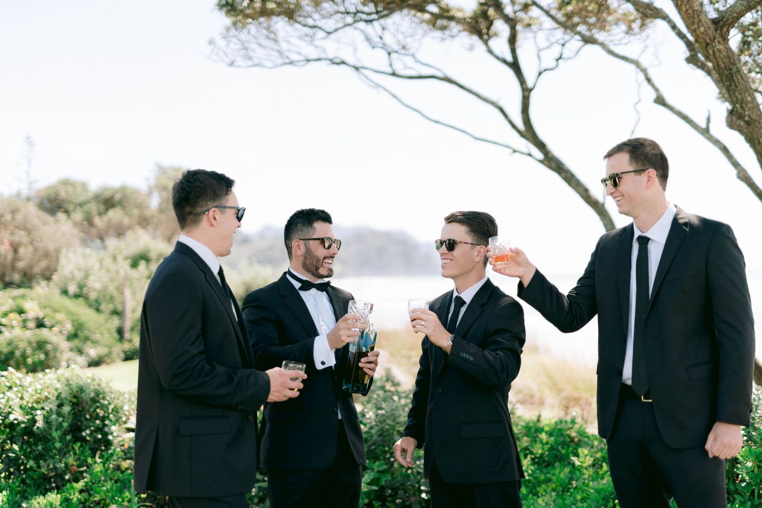 A group of groomsmen in black suits laugh and share drinks together outdoors during a wedding celebration.