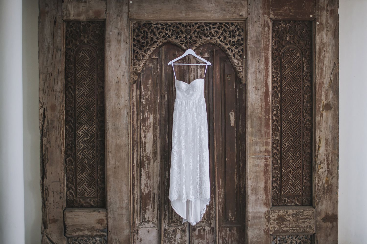 A white lace wedding dress hangs on a wooden hanger against an ornate vintage wooden door with detailed carvings.