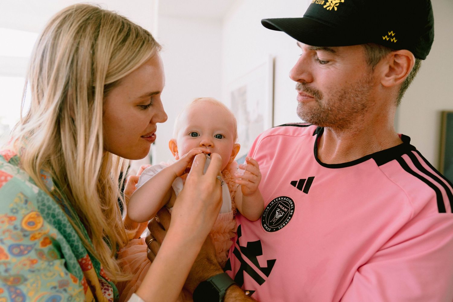 Two adults in a tender moment feeding their infant baby together at home while wearing casual attire.