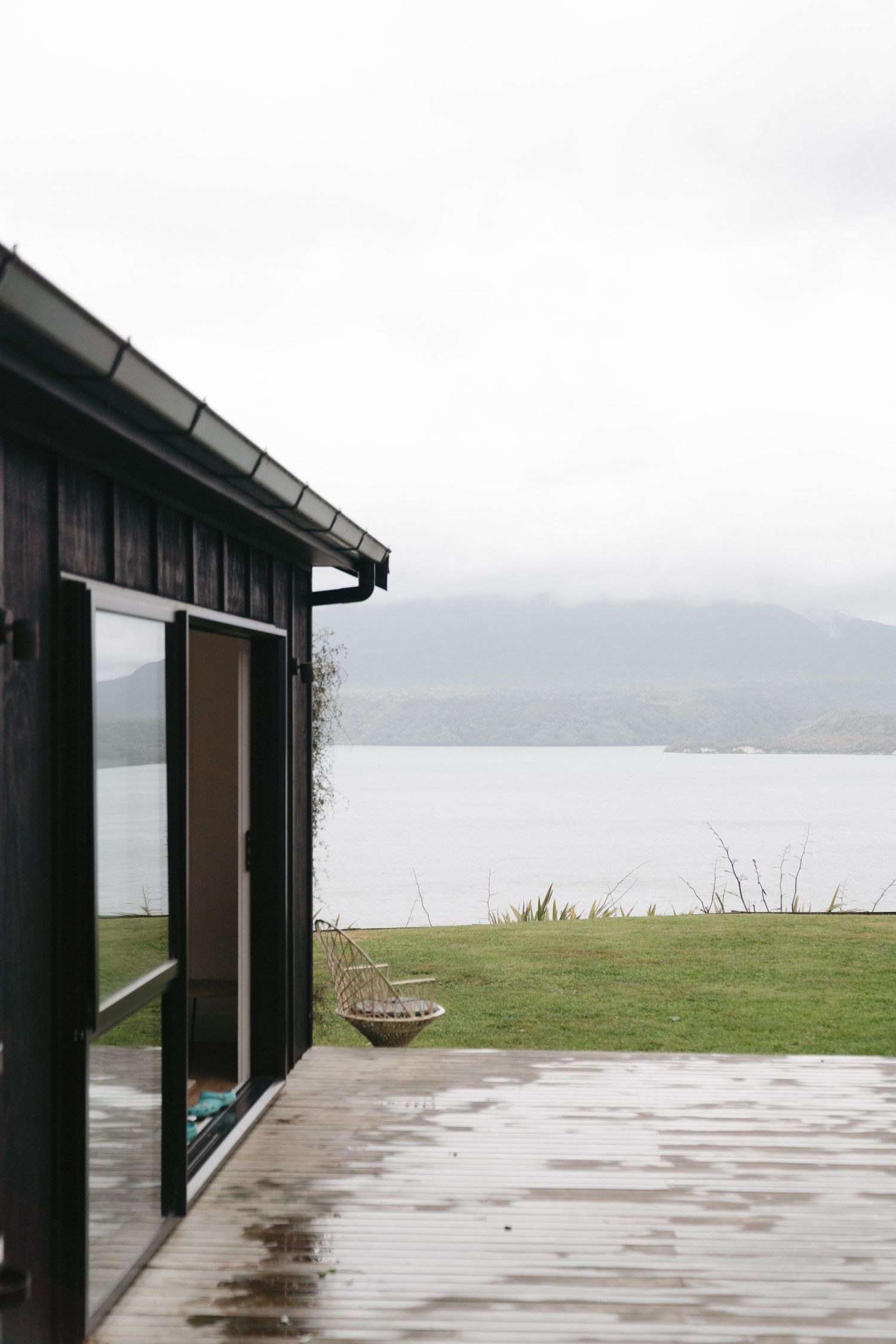Modern black cabin with deck overlooking a misty lake and mountain landscape.
