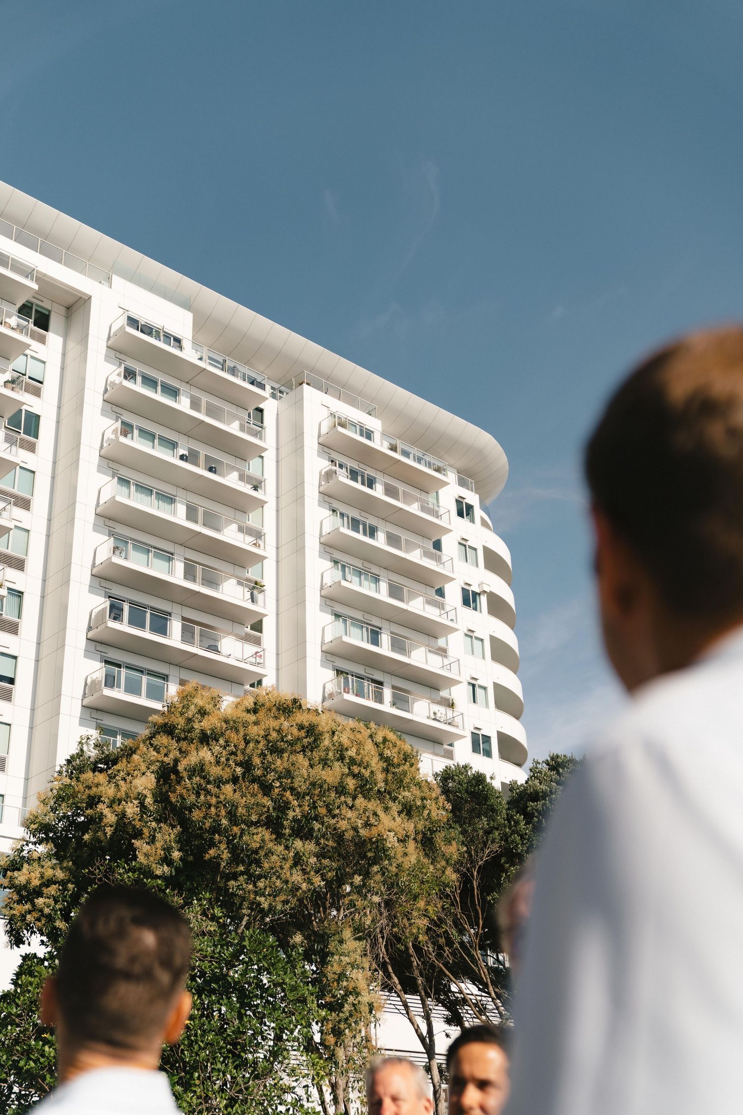 Modern white apartment buildings with balconies behind a group standing outdoors.