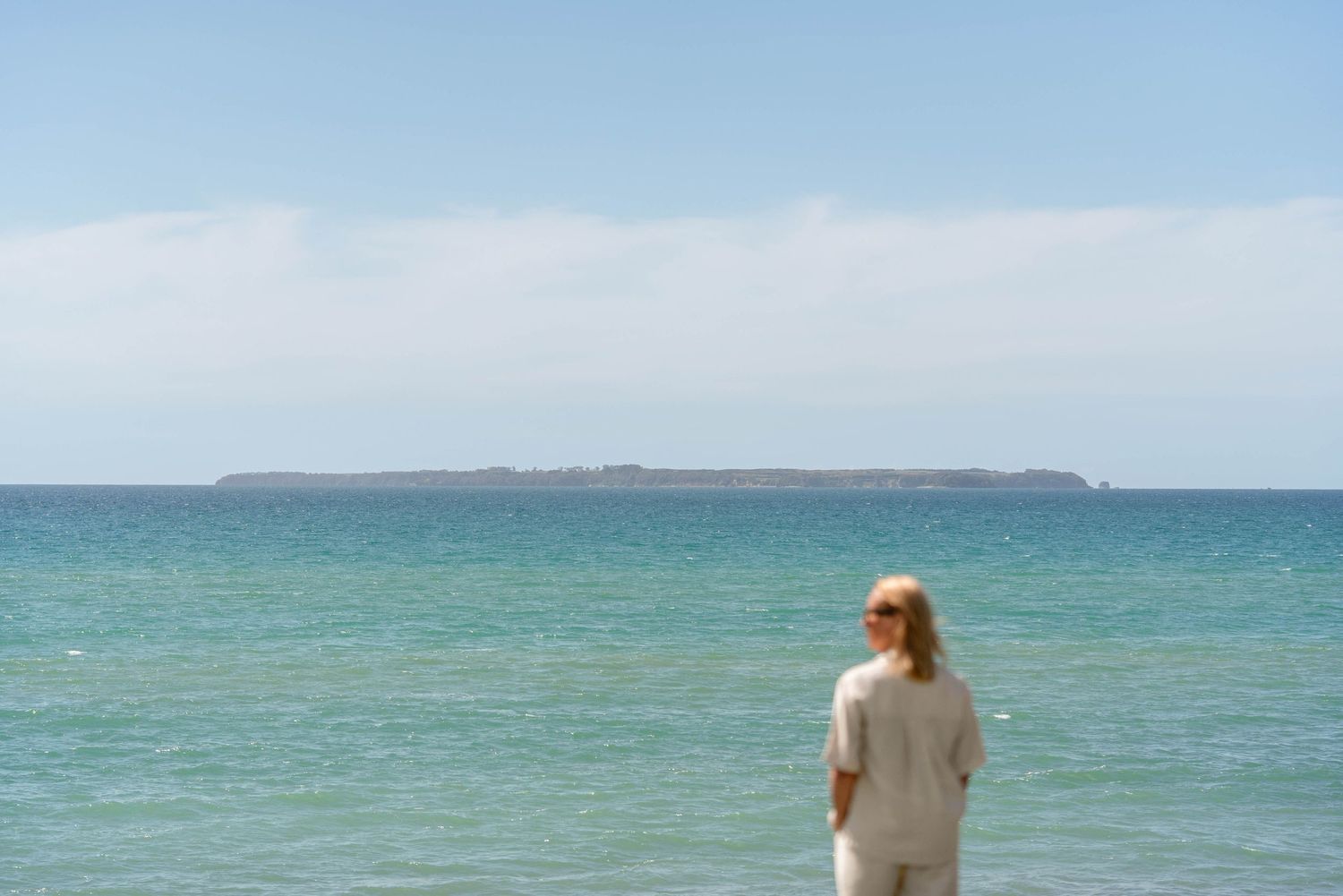 A person gazes at the turquoise ocean horizon on a sunny day with distant land visible across the water.