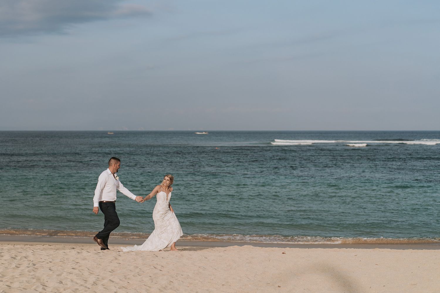 A couple in wedding attire walks along a serene beach with rolling waves and cloudy skies in the background.